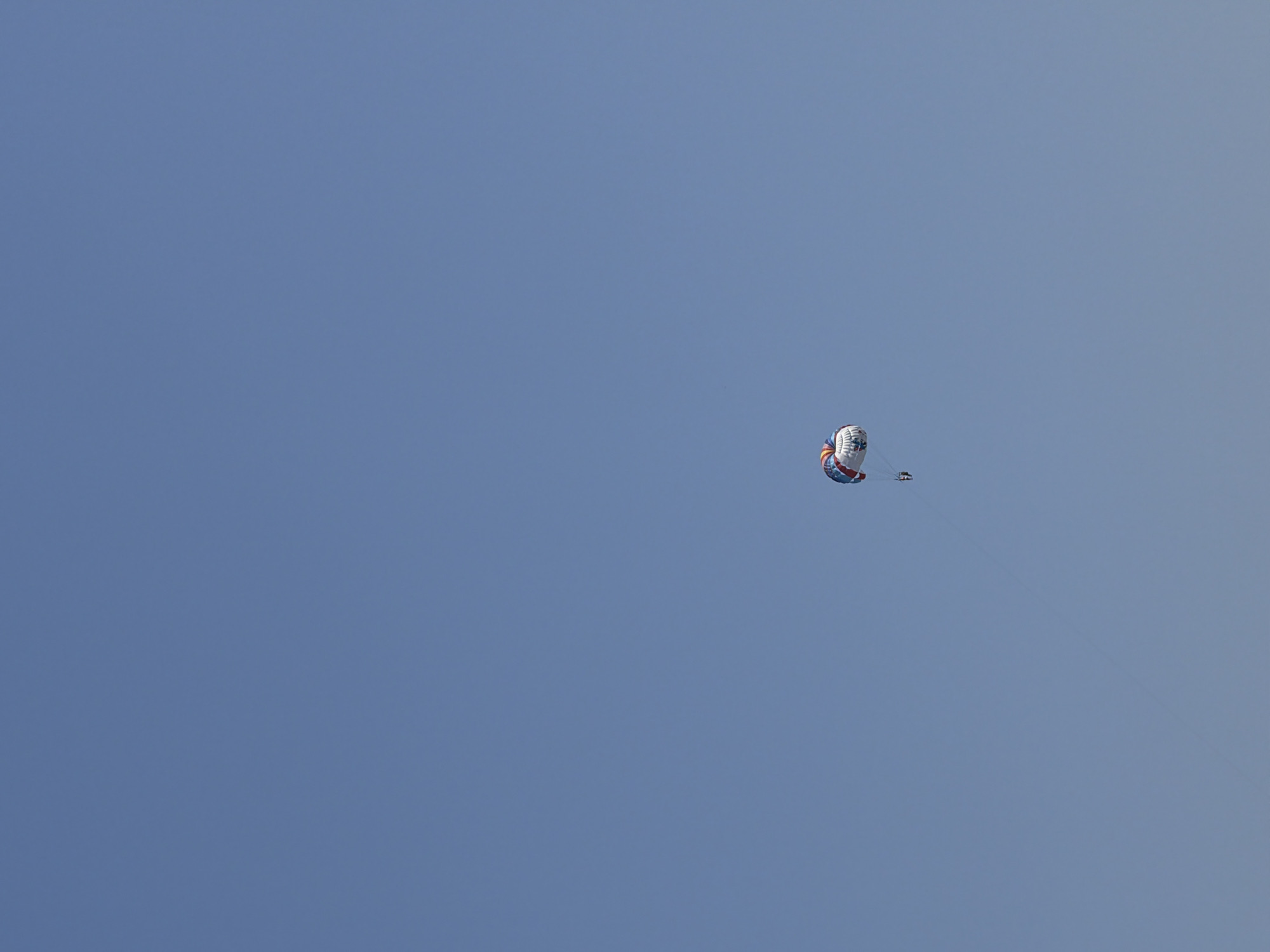 A parasailer is suspended in a clear, bright blue sky. The parasail itself is colorful, with red, white, and blue segments, and appears to be a standard design for recreational use. A small figure, presumably the parasailer, is visible hanging beneath the canopy. The parasailer is attached to the canopy by ropes or lines, which extend from the parasail to the individual. The overall scene suggests a sunny day, likely in a coastal area where parasailing is a popular activity. The image does not contain any elements that specifically identify it as being in Marmaris, Turkey, such as landmarks, signage, or distinct geographical features.