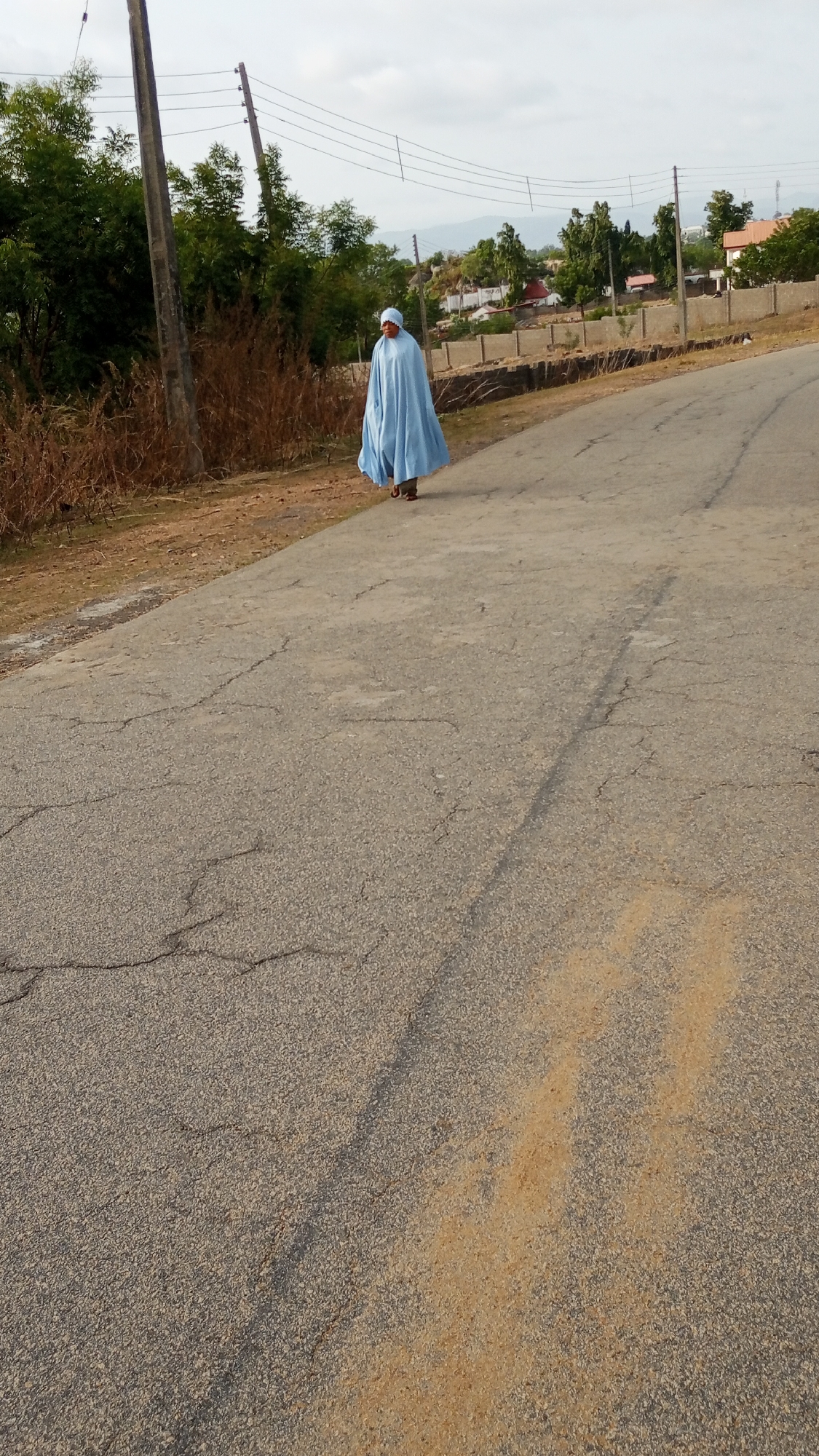 A full shot of a woman wearing a long, light blue robe and head covering is walking on a cracked gray asphalt road. The location is a road in Lokoja, Nigeria. The environment includes vegetation on the left, utility poles, and houses in the background. The weather appears clear and bright.