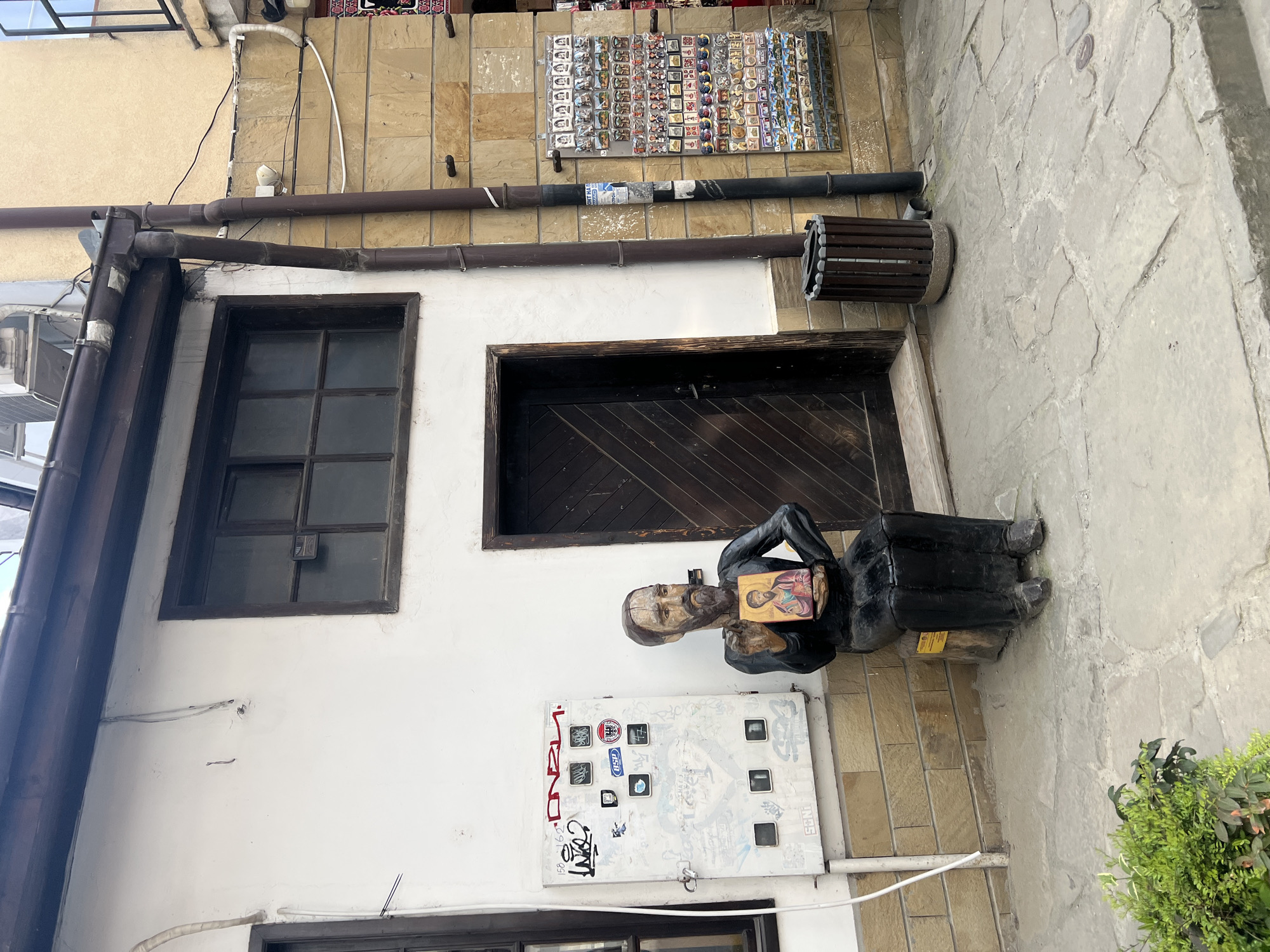 The image captures a detailed view of a narrow, stone-paved alley or courtyard in Veliko Tarnovo, Bulgaria, under clear daytime conditions.
The central subject is a life-sized, dark-robed statue of a bearded man, possibly a monk or religious figure, seated to the left of a dark wooden door. He holds an ornate religious icon, likely Orthodox, depicting two stylized figures.
The buildings exhibit a mix of architectural elements: white plastered walls, light-brown stone or brick sections, and dark-framed wooden windows and doors. Above the statue's immediate left, a large, dark-framed window with multiple panes is visible. Several brown and dark-colored drainage pipes and utility conduits run along the building facades, with a segment of one pipe showing faint, illegible text or labels. A slatted wooden waste bin is affixed to the wall to the right of the door.
Above the door, a display board showcases numerous small, rectangular items, presumably souvenirs such as magnets or badges. Below the statue, a white electrical box or panel is covered in various stickers and graffiti. Visible text on these stickers includes "ALBANIA" featuring an eagle emblem, "TSA," "FSA," and repeated Arabic script, possibly "محمد" (Muhammad). Other graffiti includes "NGS" and indistinct symbols. A vibrant green potted plant sits on the uneven stone paving to the right of the statue, adding a touch of natural color to the urban setting. The scene conveys a sense of historical charm and everyday life in an old town.