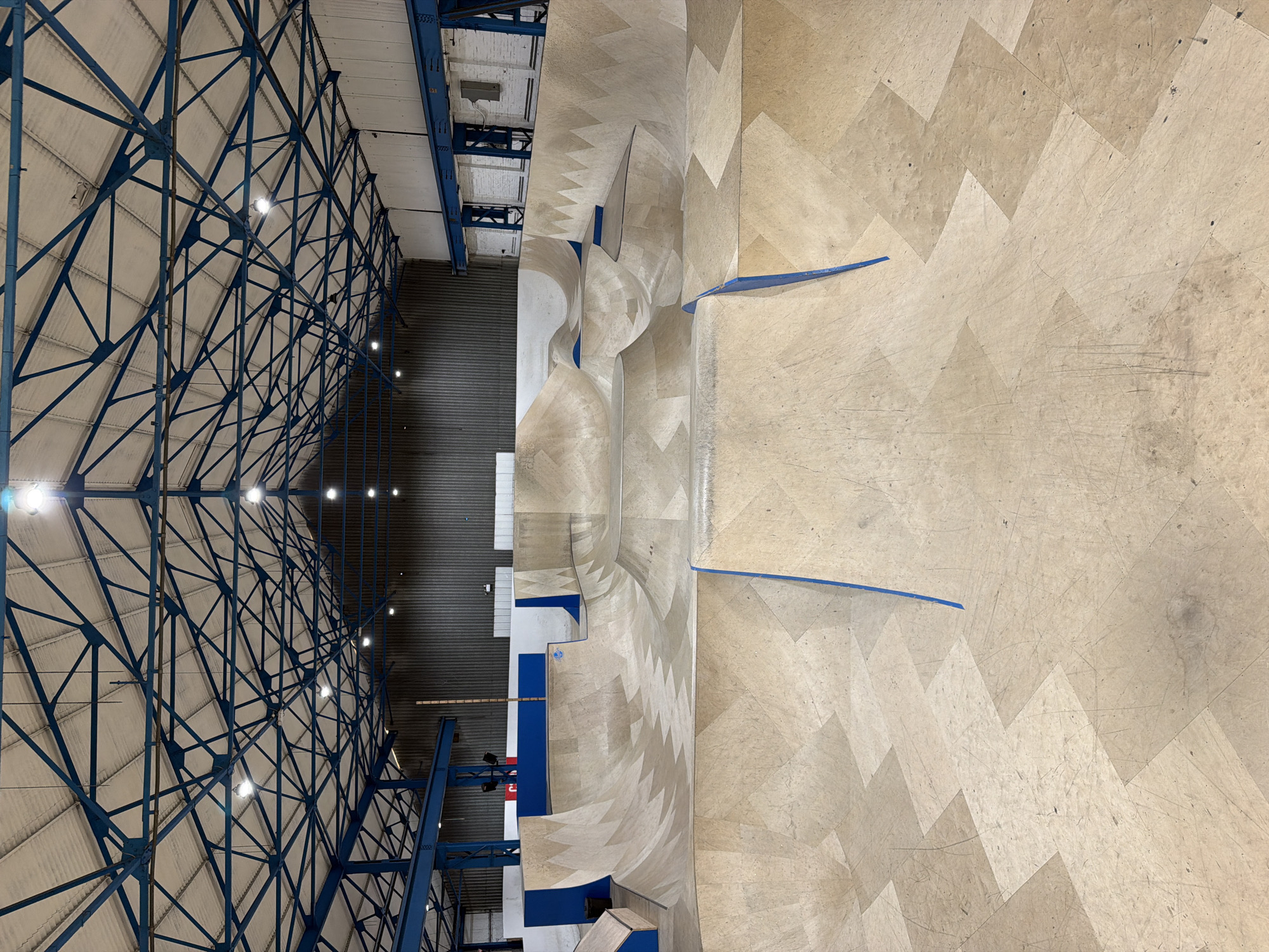 The image shows the interior of a large, indoor skatepark, specifically "The Source Park" in Ashfield, United Kingdom. The building has a high, arched roof supported by a blue, triangular lattice framework. Rows of bright, round light fixtures are attached to this framework. The skatepark floor is made of light-colored wood panels arranged in a herringbone pattern. A prominent feature of the park is a complex arrangement of ramps and curves designed for skateboarding and BMX biking. These structures are primarily light-colored wood, with blue accents on the edges. The park appears to be empty of people.