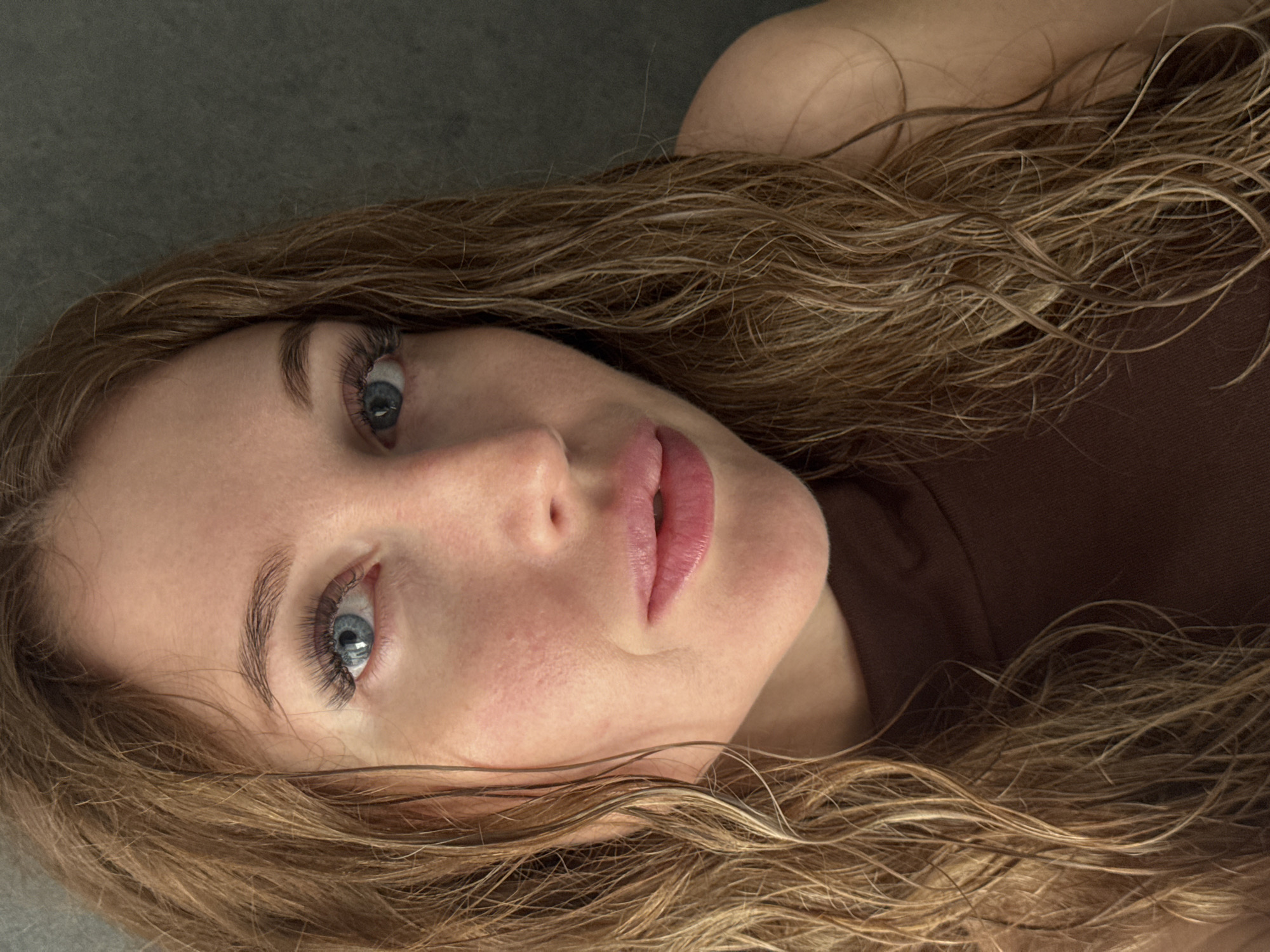 A close-up, head-and-shoulders selfie is captured in the city of Chaikovsky, Russia. The subject, a woman with light skin and light brown, wet hair, is looking directly at the camera. Her eyes are blue, and she is wearing false eyelashes. Her lips are slightly parted and appear to be tinted pink. She is wearing a brown, sleeveless top. The background is a plain, dark gray surface. The lighting suggests the photo was taken indoors, possibly in a bathroom or bedroom, with natural light coming from the side. There is no indication of the specific location within Chaikovsky beyond it being an indoor setting.