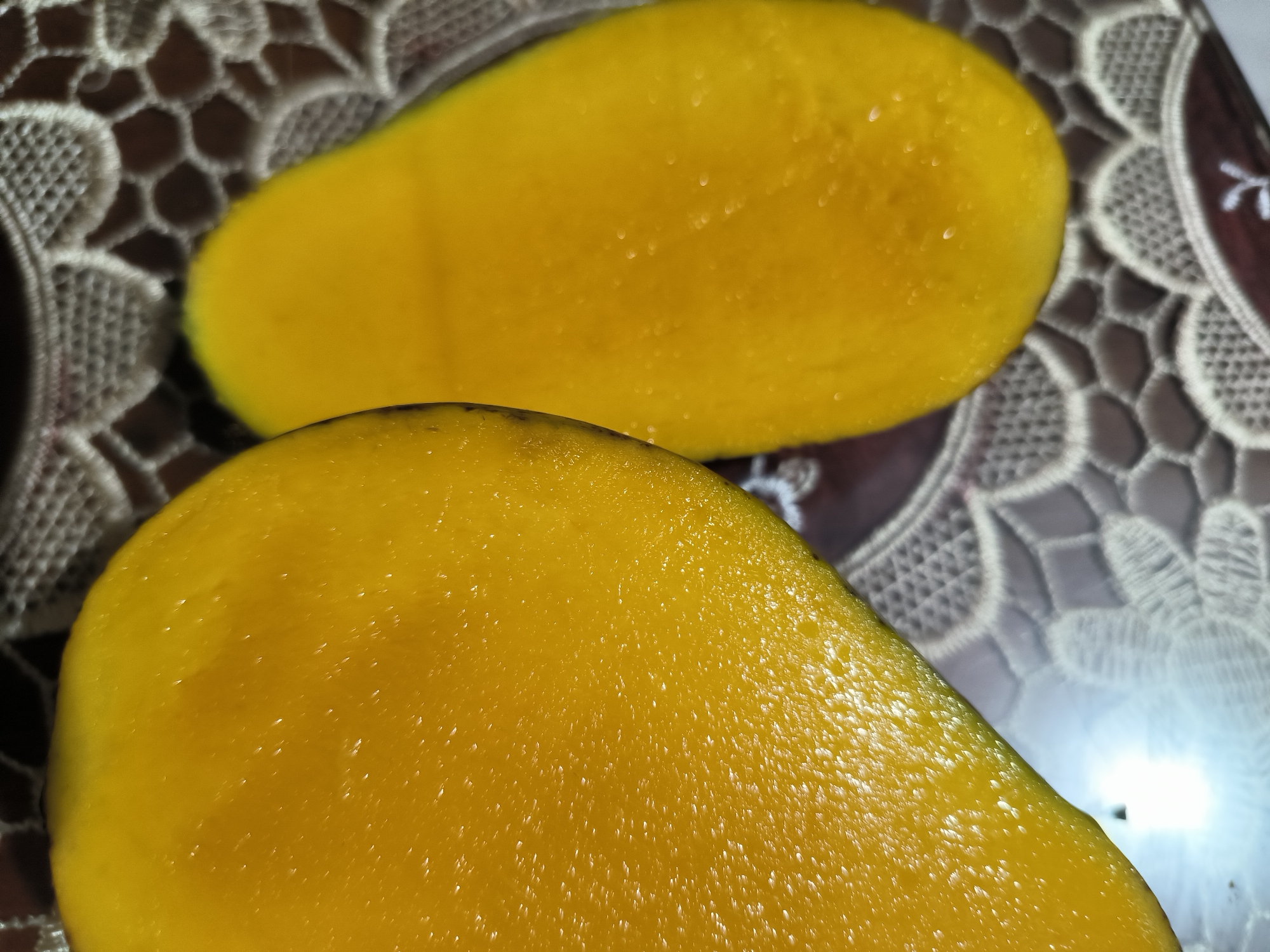 The image shows two halves of a mango fruit, cut open and placed on a surface covered with a patterned tablecloth. The mango flesh is a vibrant yellow color. The location is Lhokseumawe, Indonesia. No people are visible in the image. The subjects are the mango halves, and the object is the tablecloth. The action is the display of the fruit. No interactions are evident. The environment consists of the immediate surroundings of the fruit and tablecloth.