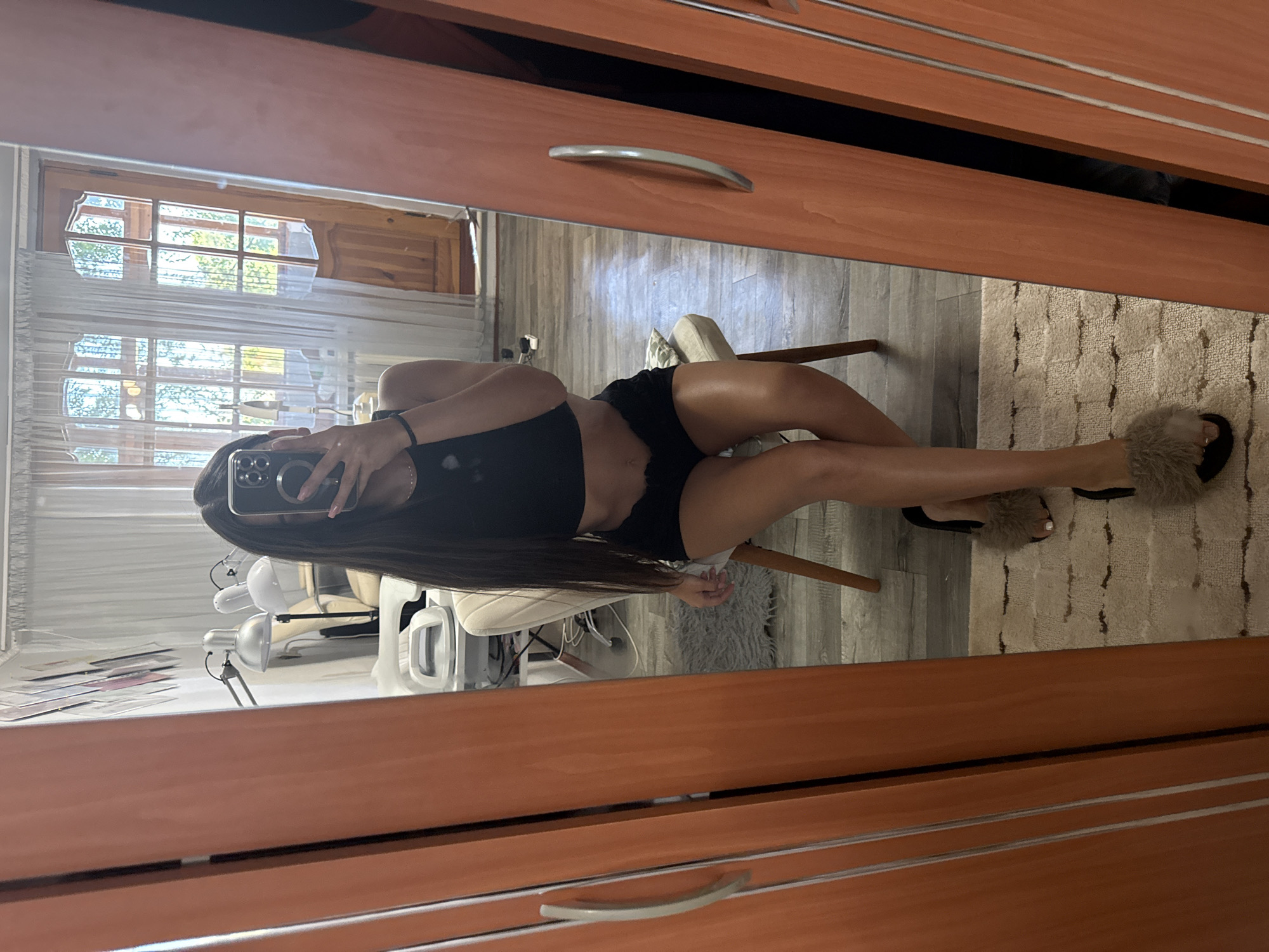A young woman with long, dark hair is taking a mirror selfie in what appears to be a room with a wooden wardrobe and a window with sheer curtains. She is wearing a black crop top and black shorts, and furry slippers. The room has a wooden floor and a rug. In the background, there are some items that suggest a beauty or nail salon, with lamps and equipment visible. The window shows trees and a wooden door. The overall impression is of a personal space, likely a home, with elements of a professional setting integrated.