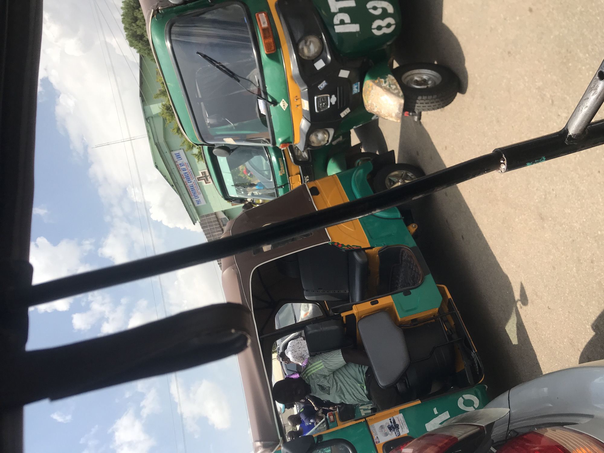 In a street scene in Jos, Nigeria, three auto-rickshaws, known locally as "Keke Napep," are visible, parked on a paved road under a cloudy sky. The auto-rickshaws are predominantly green and yellow. The foremost auto-rickshaw has the number "89" painted in white on its front. A man wearing a green and black striped t-shirt is seated in the driver's position of the second auto-rickshaw from the front. The auto-rickshaw on the left, partially obscured by a silver vehicle's rear, is also visible. In the background, a light green building with the text "THE CATHEDRAL CHURCH OF ST. LUKE" is present, along with trees and overhead wires. The scene appears to capture a moment in the bustling urban environment of Jos.