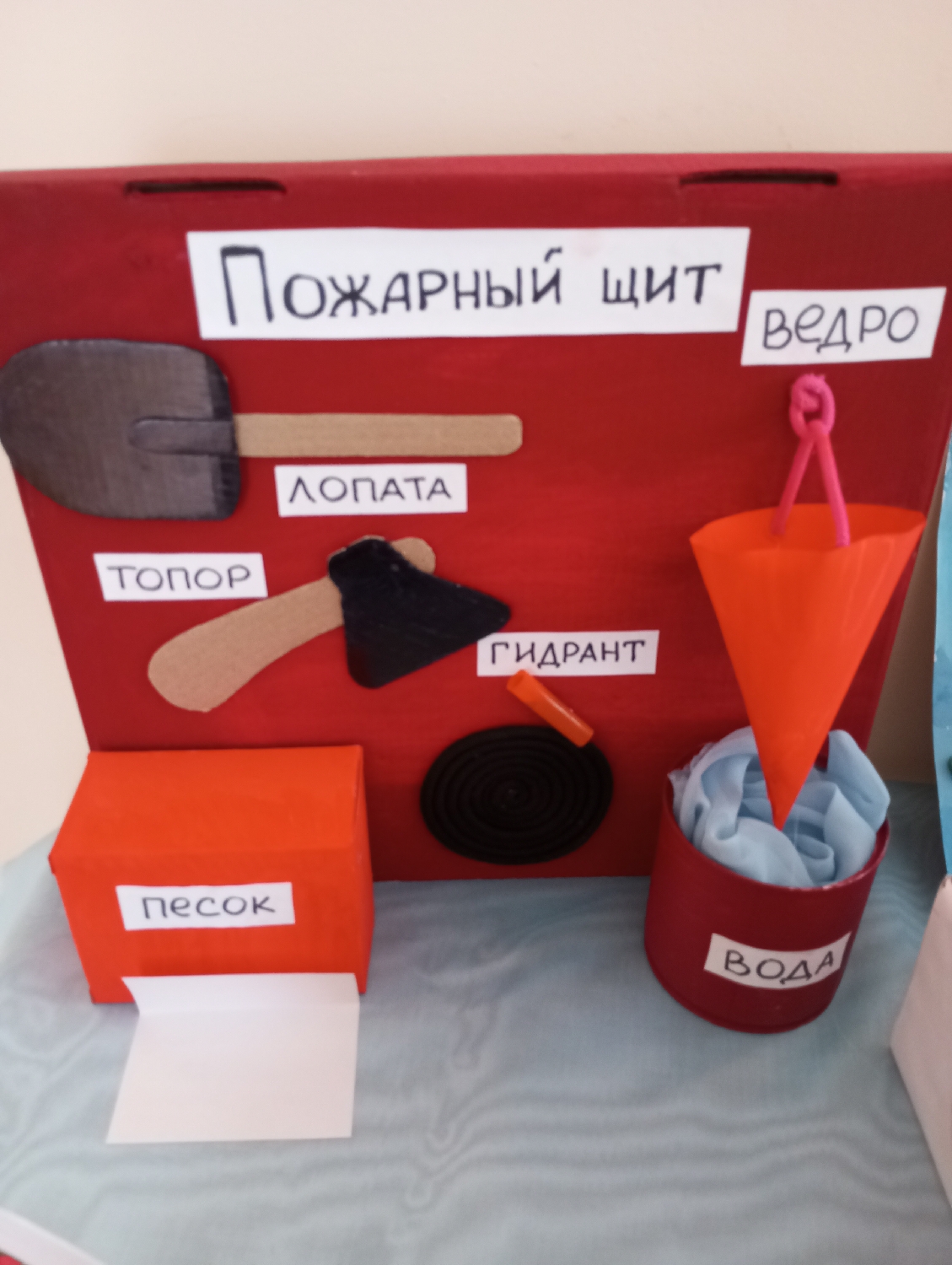 This image captures a vibrant, handmade educational display illustrating firefighting tools, likely designed for children. The central element is a red rectangular board, resembling a "fire shield" (Пожарный щит), as indicated by the prominent Russian text at the top.
Affixed to this red board are simplified, two-dimensional representations of various firefighting equipment, each clearly labeled in Russian:
* A shovel, with a grey blade and a brown handle, is labeled "ЛОПАТА" (shovel).
* Below it, an axe with a black head and a brown handle is labeled "ТОПОР" (axe).
* On the right side of the board, a black coiled hose with a small orange nozzle represents a "ГИДРАНТ" (hydrant).
* Above the hydrant, an orange cone-shaped object with a pink rope loop serves as a "ВЕДРО" (bucket).
Arranged on the light blue patterned surface in front of the main board are two additional items:
* An orange box, from which a strip of white paper emerges, is labeled "ПЕСОК" (sand), signifying a sand box used for fire suppression.
* Next to it, a red cylindrical container filled with light blue fabric is labeled "ВОДА" (water), representing a water supply.
The display is meticulously crafted, suggesting its use in an educational context such as a classroom or a safety awareness program. The clear, block-lettered labels in Russian facilitate understanding for learners. The scene is static, set indoors, with no people visible. The craft's nature points to an informational or instructional purpose, possibly in a school or kindergarten in Izhevsk, Russia.