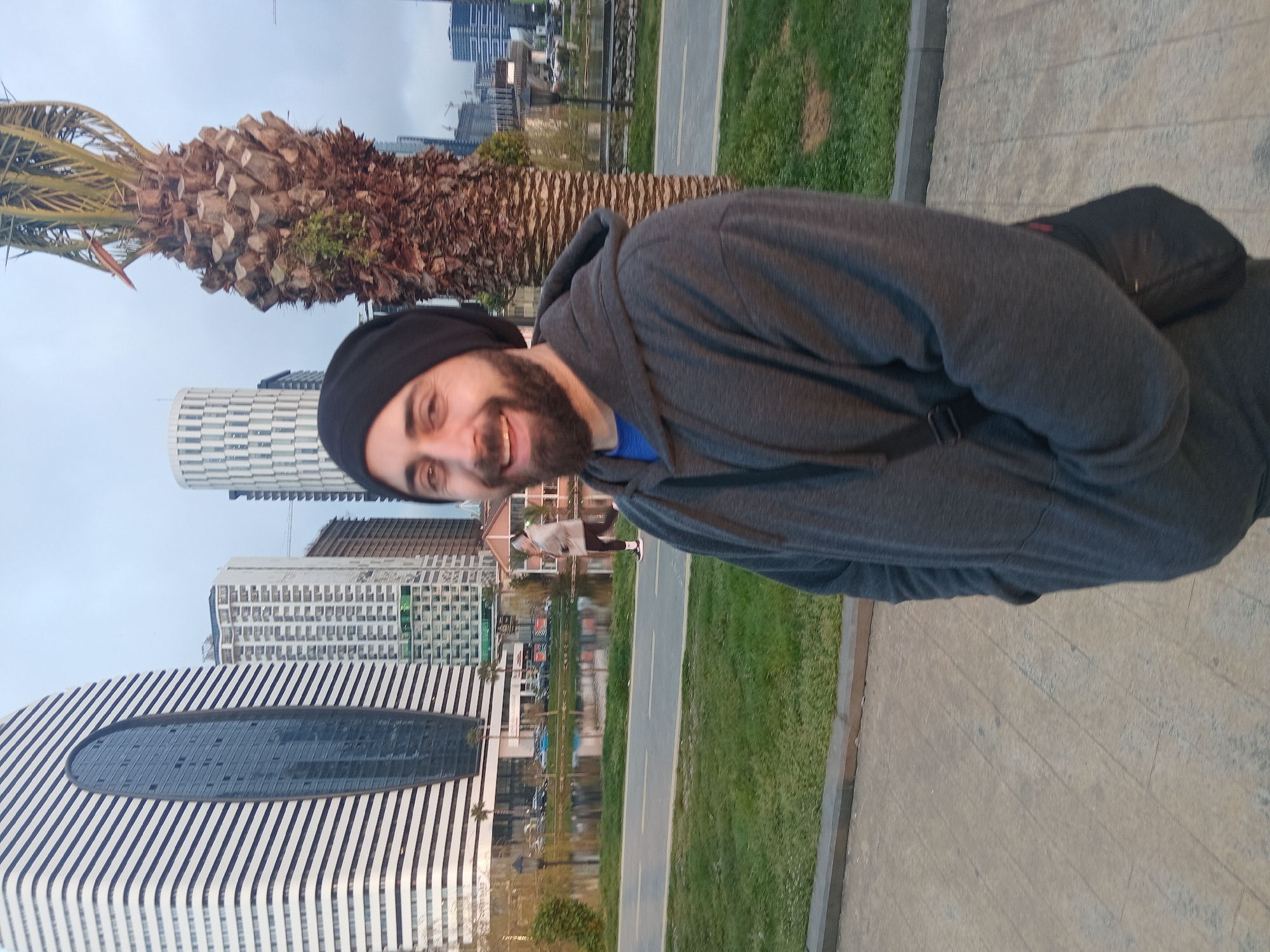 A man with a dark beard and wearing a black beanie and gray hoodie stands on a paved area in Batumi, Georgia. The man is smiling and appears to be taking a selfie. Behind him is a line of trees and green space. In the background, there is a reflective body of water, and modern architectural buildings including the white building with a diagonal black stripe design, which is located on Sherif Khimshiashvili Street in Batumi. There is also a palm tree in the background. The overall lighting suggests it is daytime.