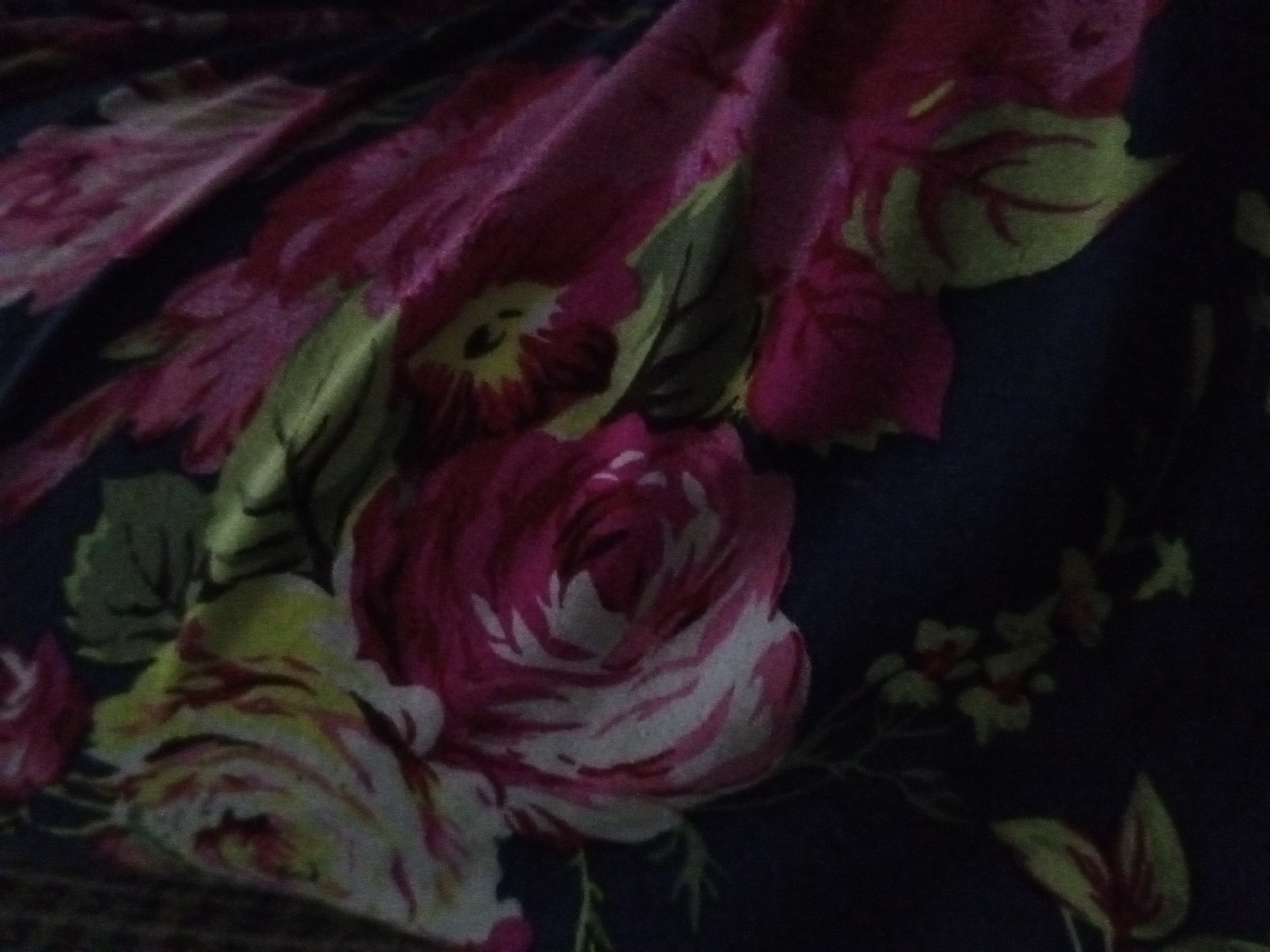 The image depicts a close-up view of a floral-patterned fabric. The dominant colors are shades of pink, white, green, and a dark blue background. The fabric is bunched or folded, creating a sense of depth and texture. Large roses are the main floral motif, with smaller floral patterns scattered throughout the fabric. The lighting is subdued, resulting in some areas of the fabric appearing darker than others. There is no indication of the location being Tinaquillo, Venezuela.