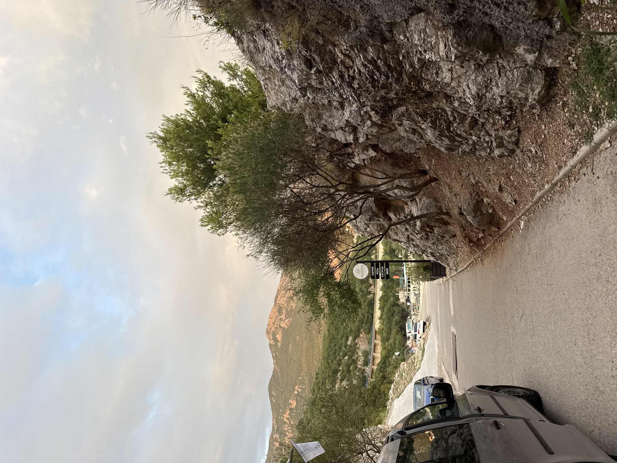 The image depicts a scenic road winding down a hillside in Kamenovo, Montenegro. The road is paved and curves to the right, with a steep, rocky cliff face on the left and a lush, green hillside on the right. A few trees with sparse foliage grow along the cliff's edge.
Several vehicles are parked or driving on the road, including a dark-colored SUV in the foreground and a blue car further down. In the background, a small village or resort area is visible, with buildings nestled amongst the greenery. A signpost with text is also visible, indicating "COFFEE PIZZA COCKTAIL BEER". A white flag with black text, possibly a banner or sign, is tied to a railing near the foreground vehicles.
The sky is overcast with scattered clouds, suggesting it might be late afternoon or early evening. The overall mood is serene and picturesque, capturing the natural beauty of the Montenegrin coast. The image appears to be taken from a slightly elevated perspective, looking down the road.