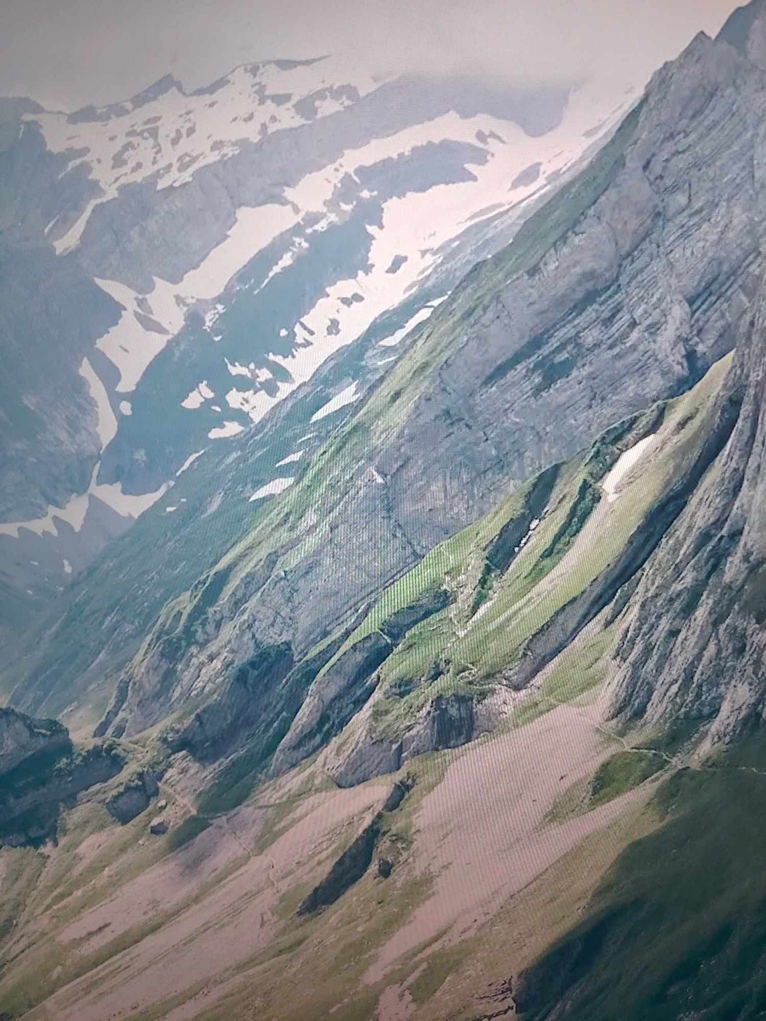 The image depicts a mountainous landscape. The mountains exhibit steep, layered rock formations with patches of snow at higher elevations. The lower slopes are covered in green vegetation, transitioning to exposed rock and gravelly terrain. A narrow trail or path is visible winding along the slope. The scene appears to be set in a remote, rugged environment. Location details (city of Unknown, Unknown) are not reflected in the image.