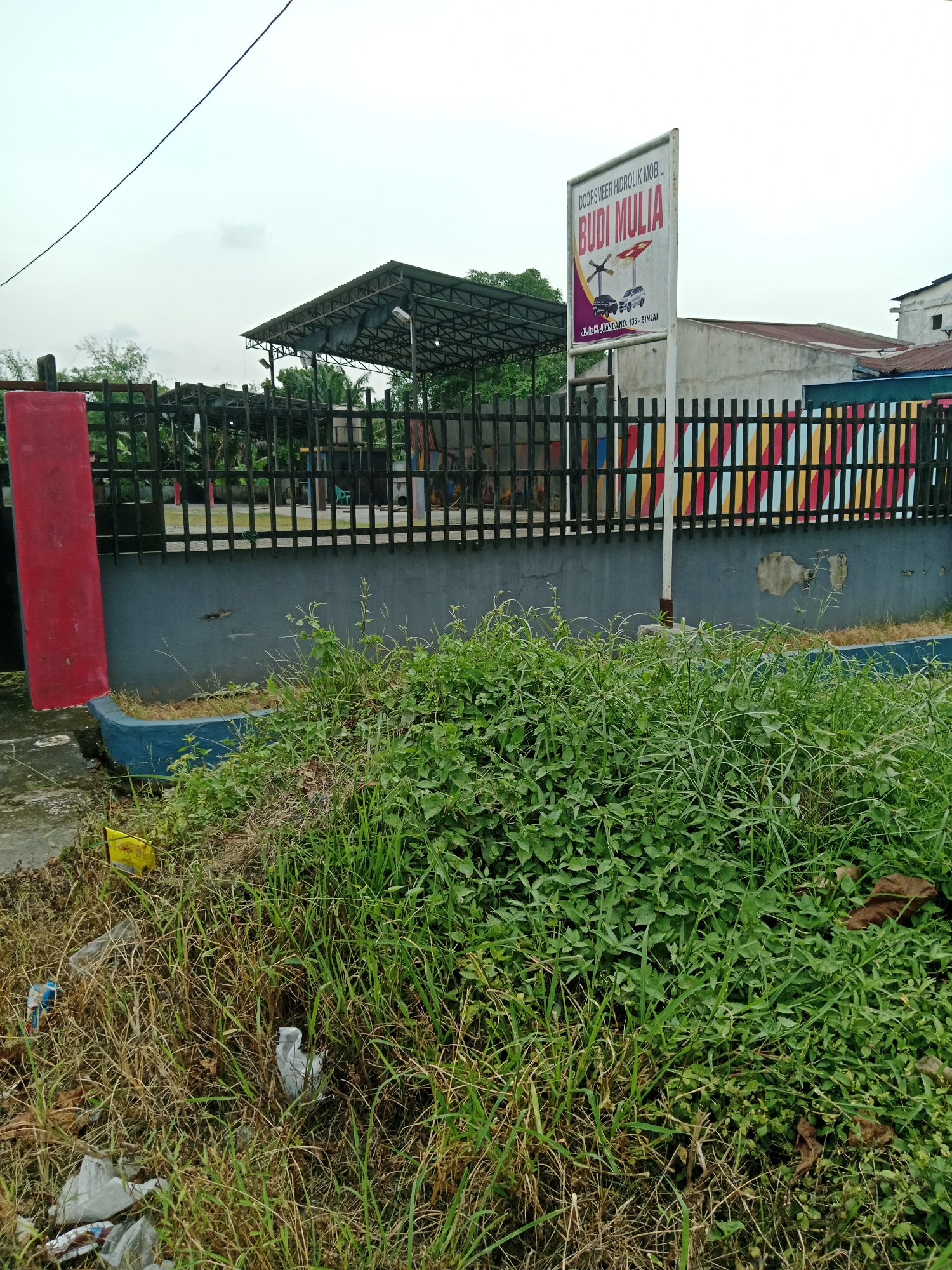 The image depicts an outdoor scene, likely in the City of Binjai, Indonesia, featuring a car service establishment. In the foreground, overgrown grass and weeds are visible, with some scattered litter including plastic bags and bottles. A red rectangular structure is on the left side of the image, adjacent to a gray concrete wall. A black metal fence runs along the wall, which is decorated with colorful diagonal stripes of red, yellow, green, and blue. Behind the fence, a covered structure with a metal roof, likely a service bay, is visible. A white signpost with text and images is prominently displayed on the right side of the fence. The sign has text that reads "DOOR SMEER HIDROLIK MOBIL BUDI MULIA" and displays illustrations of a car and a lifting mechanism. Below this, the address "JL. RA. JUANDA NO. 135 - BINJAI" is printed. In the background, a building with a reddish-brown roof and a white facade can be seen, along with more vegetation and utility poles. The sky is overcast.