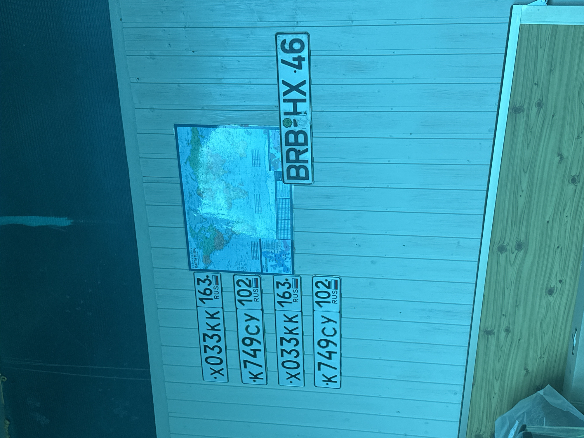The image shows a wall with several Russian license plates and a world map poster. The license plates are arranged in two columns of two plates each, with a single license plate to the right. The license plates appear to be from a Russian city, indicated by the "RUS" designation on each plate. The world map poster is mounted on the wall above the license plates. The overall lighting in the image has a strong blue tint. The location is described as being in Novokuybyshevsk, Russia, which is a city in Samara Oblast, Russia. The license plates themselves provide further clues. The numbers "163" and "102" on the plates likely refer to the regional codes for Samara Oblast and Orenburg Oblast, respectively, both of which are neighboring regions to Novokuybyshevsk. The license plate to the right, "BRB HX 46", also displays a regional code "46", which corresponds to Kursk Oblast, another region in Russia. The context suggests these plates may be decorative items or souvenirs collected from various regions.