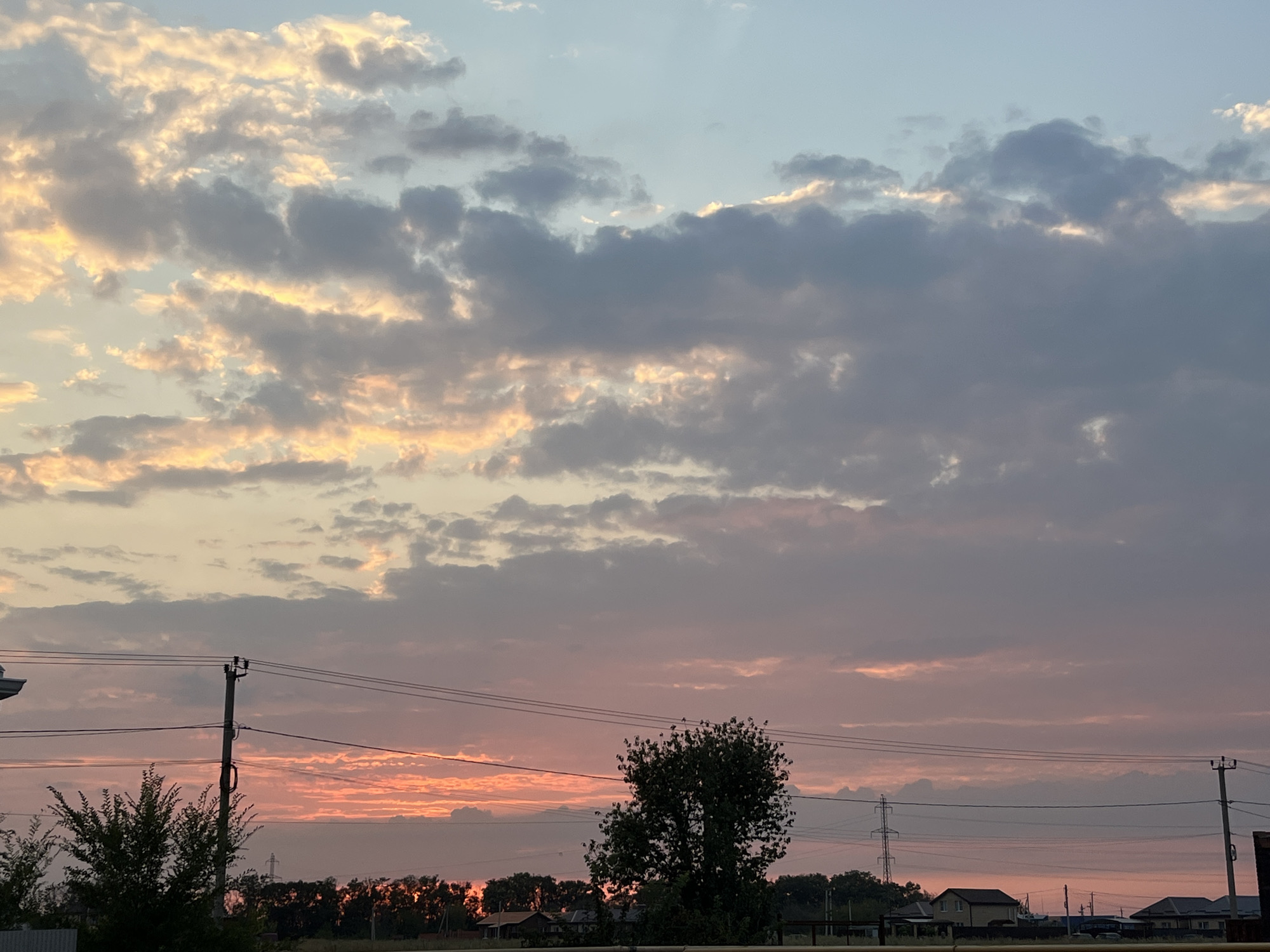 The image depicts a rural or suburban landscape in the Taganrog City District, Russia, under a twilight sky. The sky is filled with a dramatic display of clouds, illuminated by the setting sun, casting hues of orange, pink, and gold against a backdrop of soft blue and gray. In the foreground, power lines crisscross the scene, supported by utility poles, indicating infrastructure common in such areas. A prominent tree stands in the middle ground, silhouetted against the vibrant sky. Beyond the tree, a line of buildings and trees forms the horizon, suggesting a small settlement or rural community. The overall impression is one of a serene, yet visually striking, evening in the Russian countryside.