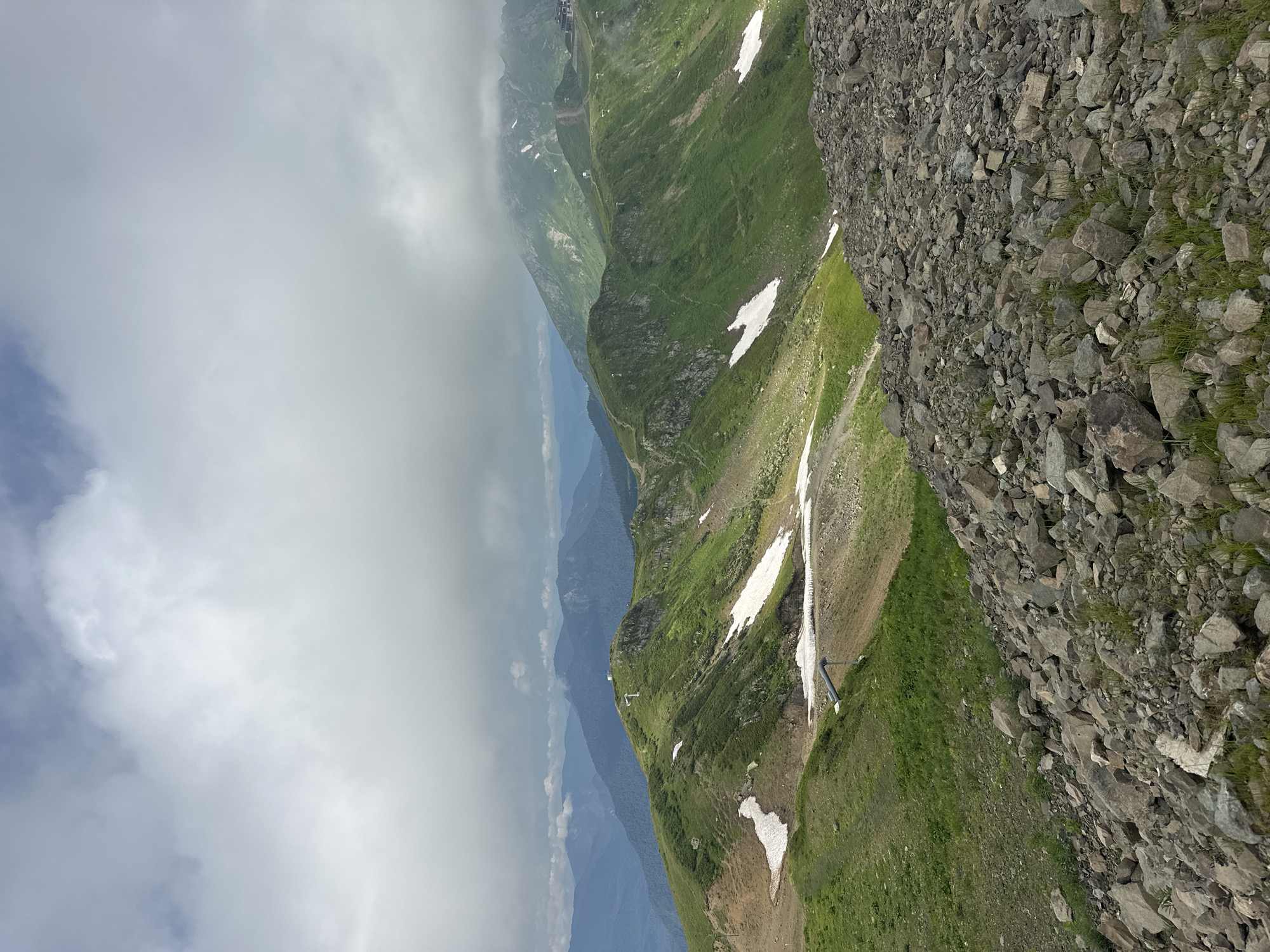 A wide-angle, vertical, eye-level shot captures a mountainous landscape under a cloudy sky. The foreground is dominated by a rocky slope strewn with various grey and brown stones of different sizes, interspersed with sparse green grass. Beyond the rocky foreground, rolling green hills descend, marked by patches of melting white snow. A faint path or road winds through the grassy slopes. In the middle ground, more green, rounded mountains rise, some with visible snow remnants. A small white structure with an antenna stands atop one of the peaks. Further in the distance, layers of hazy blue mountains recede into the horizon, partially obscured by a thick layer of grey clouds that fill the upper portion of the frame. The overall scene depicts a rugged, natural environment.