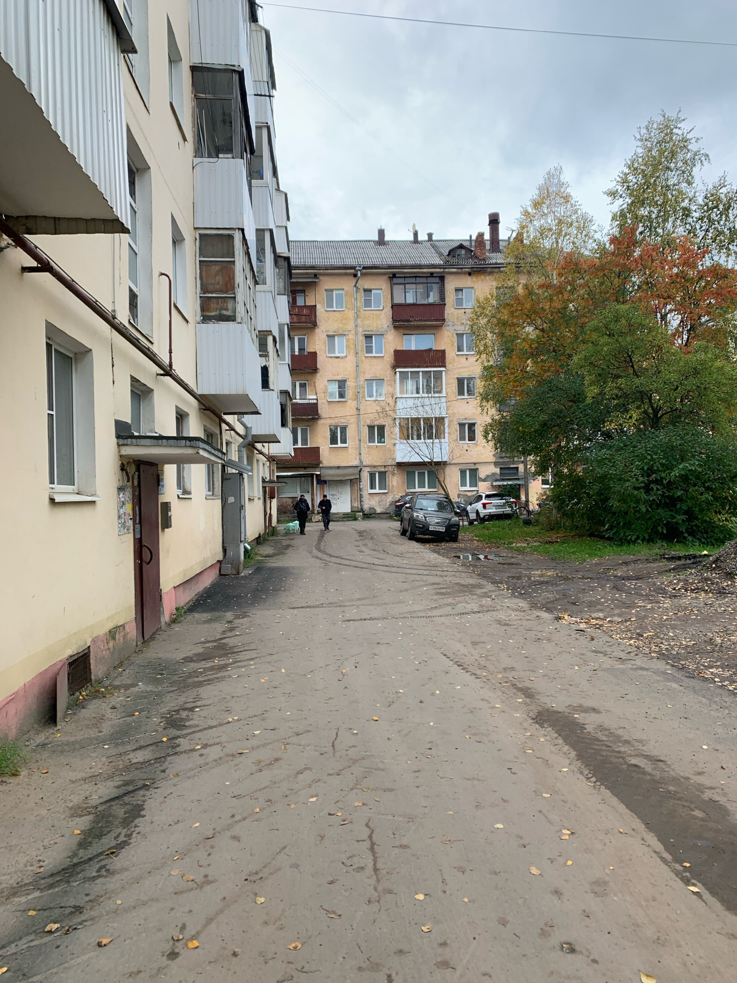 The image shows an exterior view of an apartment building in Arkhangelsk, Russia. The building is light yellow with multiple balconies, some enclosed and some open. A taller apartment building, also yellow, is visible in the background with a grey tiled roof. The area in front of the buildings is a paved courtyard with some puddles and fallen leaves, indicating an autumn setting. Two individuals are walking away from the camera down the courtyard path. Several cars are parked in the courtyard, including a dark SUV on the left and a white SUV to the right of it. Trees with autumn foliage are present in the background. The sky is overcast.