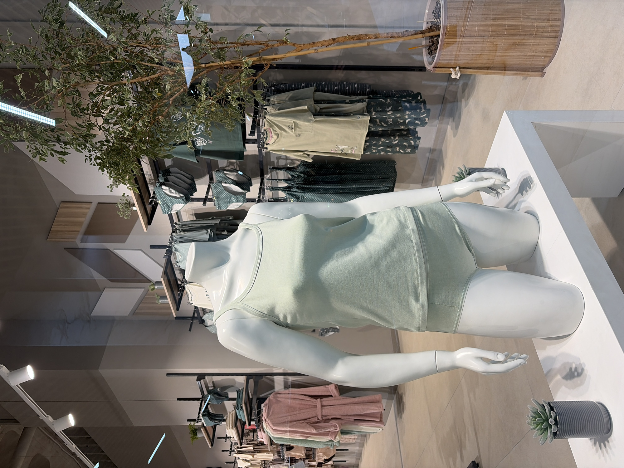 A mannequin wearing a light green tank top and matching bottoms stands on a white platform in a store display. The display is located in a retail environment, possibly a clothing store, with racks of clothes visible in the background. Various items of clothing in shades of green and pink are displayed on the racks. A potted tree stands behind the display. A small potted succulent is also placed on the platform. The location is Izhevsk, Russia.