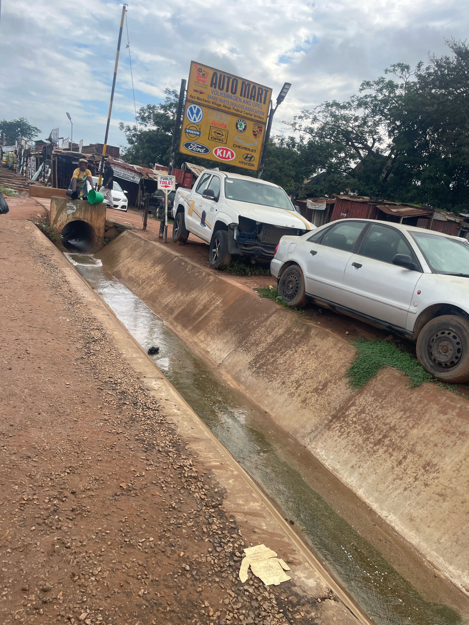 In Abuja, Nigeria, near Apo Mechanic Village, a wide concrete drainage ditch runs alongside a dirt road. The ditch contains murky water and debris, including a piece of light-colored fabric resembling a shirt lying on the bank. On the road, several vehicles are parked or abandoned. To the right, a silver sedan is parked facing away, its wheels resting on the edge of the ditch. Further back and to the left, a white pickup truck is parked. A large yellow sign with "AUTO MART" and various car logos is visible in the background, along with a public toilet sign. Two people are near the entrance of the ditch, one holding a green bucket and the other standing. A striped pole, possibly part of a barrier or signal, extends upwards. The scene depicts an area with auto repair services and a general sense of urban clutter.