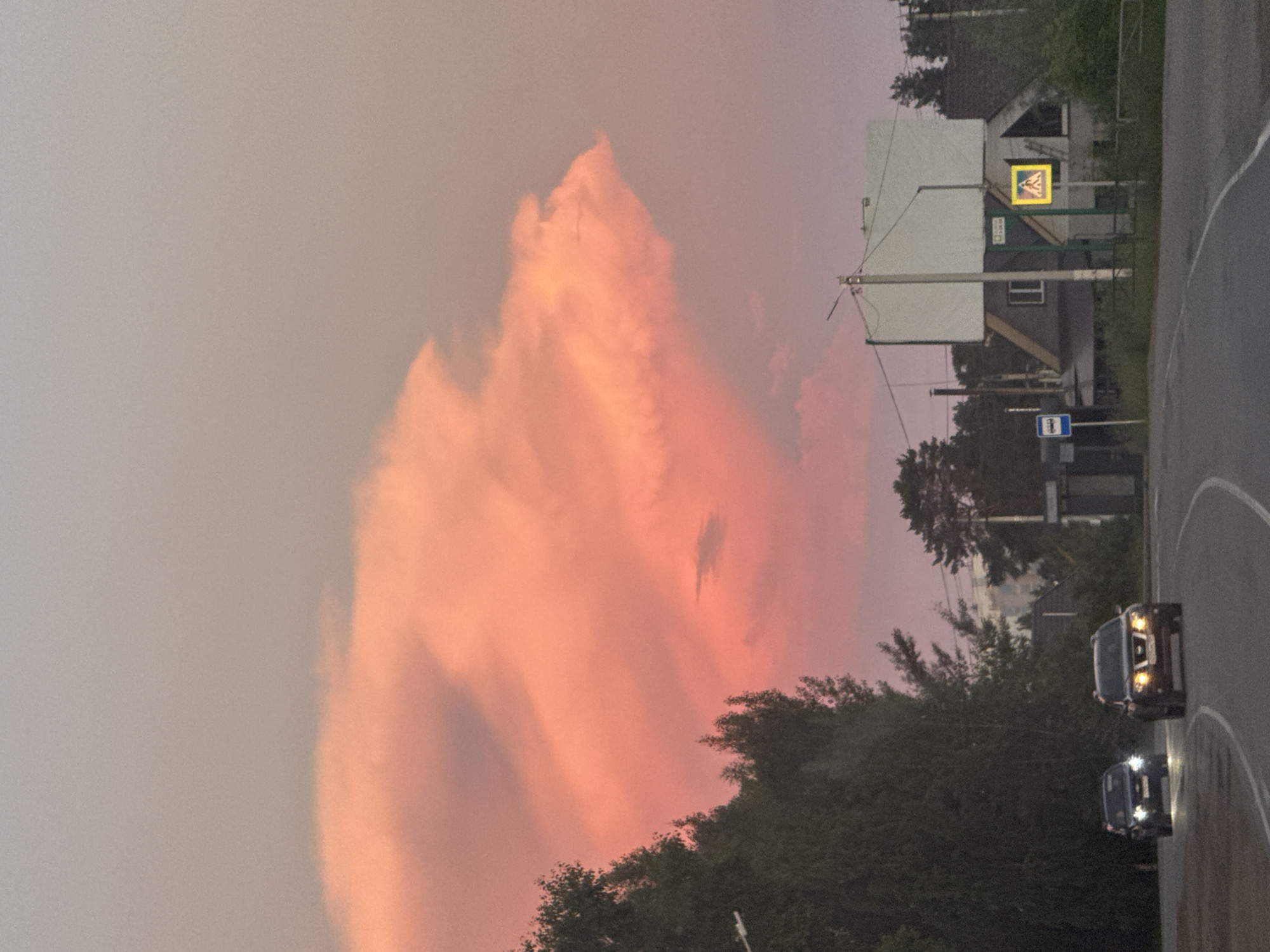 The image depicts a street scene in Chaikovsky, Russia, under a dramatic sunset sky. A large, vibrant pink cloud formation dominates the upper portion of the frame, casting a warm glow over the landscape. In the foreground, a two-lane asphalt road curves to the right. Two cars are visible on the road: a dark sedan on the left with its headlights on, and a dark SUV on the right, also with headlights illuminated.
To the left of the road, a dense line of dark green trees runs along the edge of the roadway. Behind the trees, houses with visible roofs and windows can be seen. To the right of the road, a large, rectangular billboard with a blank white surface stands prominently, mounted on poles. Adjacent to the billboard is a small, gray house with a pitched roof. A yellow pedestrian crossing sign with a black silhouette of a person is positioned in front of the house. A blue bus stop sign is also visible further down the road. The overall environment suggests a suburban or semi-rural setting within the city of Chaikovsky.