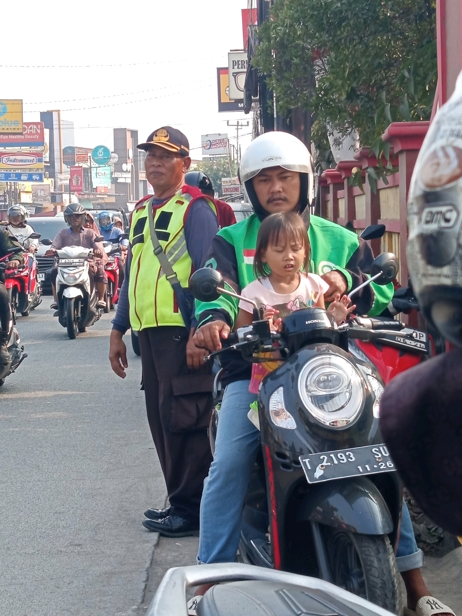 A street scene in Karawang, Indonesia, captures a moment of traffic with multiple motorbikes. In the foreground, a man wearing a white helmet and a green jacket is riding a black motorbike, with a young girl in a pink shirt sitting in front of him. Behind them, a man in a yellow vest and a dark cap stands on the side of the road, looking towards the camera. Several other motorbikes and people are visible in the background, along with various signs and buildings lining the street, indicative of a commercial area. The sky is bright and hazy, suggesting it might be daytime.
