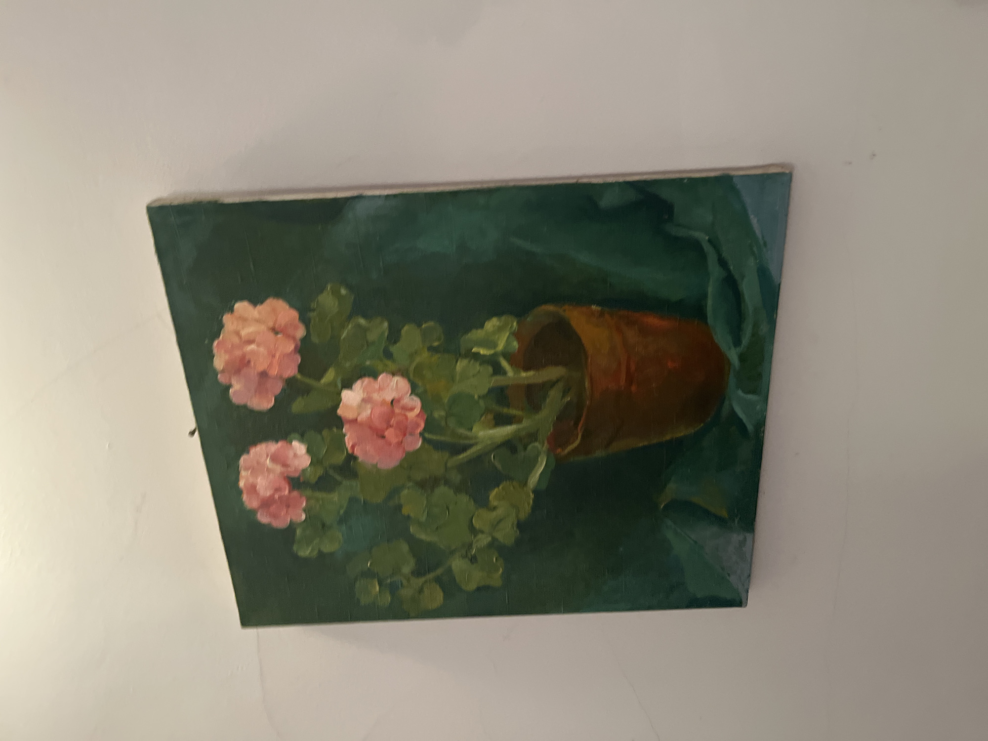 The image depicts an oil painting on canvas, likely hanging on a white wall. The painting features pink geranium flowers in a terracotta pot, depicted in a slightly tilted position. The background is dark green. The scene's location is unknown, and there's no indication it is Goce Delcev, Bulgaria. The painting appears to be an amateur work.