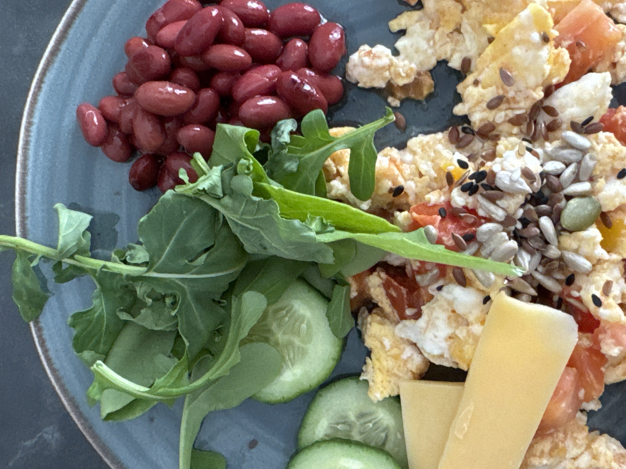 A close-up shot of a blue plate filled with food. The plate contains a mound of kidney beans, a green salad comprised of arugula and cucumber slices, scrambled eggs with what appears to be tomatoes and a generous topping of mixed seeds (sunflower, pumpkin, flax), and two slices of yellow cheese. The background is a dark, nondescript surface. There is no information to suggest that this is Chaikovsky, Russia.