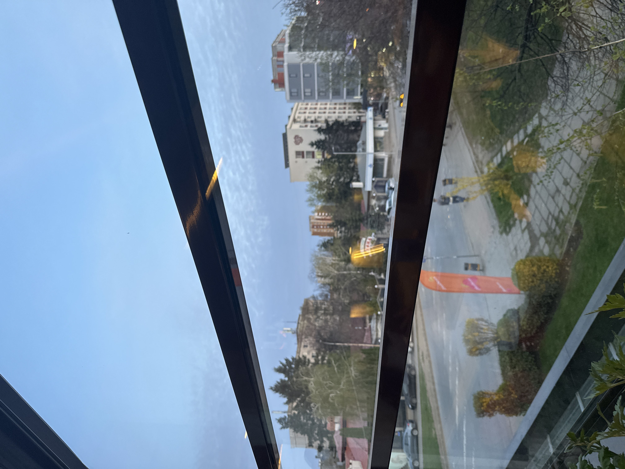 The image, originally rotated 90 degrees clockwise, depicts a vibrant urban scene in Sofia, Bulgaria, viewed from an elevated position, likely through a large window or skylight of a building. The perspective looks downwards onto a city street and surrounding architecture.
In the immediate foreground, dark metallic frames of the window structure diagonally cross the view, bordering a clear, bright light blue sky in the upper left. Some vivid green foliage, possibly from an indoor plant, is visible in the bottom right corner.
Beyond the window, the cityscape unfolds under a daytime sky with minimal cloud cover. Several multi-story buildings of varying architectural styles and colors—including white, grey, and brick-red facades—dominate the background. One prominent white building features a distinctive red heart-shaped logo on its side, with illegible text beneath it. Another building further back appears to have an antenna on its roof. A structure resembling a gas station or a small commercial building is also discernible.
A wide paved street stretches across the mid-ground, with several cars parked along its side. Abundant greenery is scattered throughout the scene, including numerous trees, some with budding green leaves and others with bare branches, interspersed with ornamental shrubs and hedges, some shaped geometrically, lining the roadside. A grassy area or park runs parallel to the street.
An eye-catching orange banner or flag stands vertically near the street, displaying a white logo that includes a distinct 'S' shape and other white, unreadable text, likely related to "Sofia" or a local event. Several yellow artificial lights, possibly from streetlights or building signs, are visible, suggesting either late afternoon or that they are illuminated during the day. The overall impression is a clear, bright day in a lively city environment. No specific human activity or identifiable people are visible.