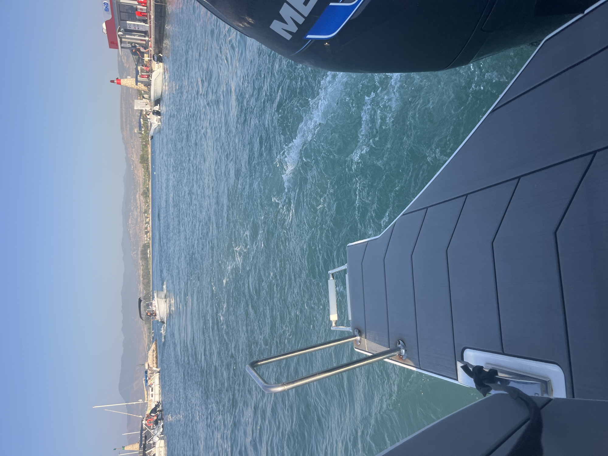 The image captures a view from the stern of a boat moving through the turquoise waters of Polis Chrysochous, Cyprus. The perspective is looking out towards a harbor area. In the mid-ground, a white motorboat with an open canopy and several occupants is visible, leaving a wake. To the left, a harbor wall is lined with other boats, including sailboats, and a lighthouse-like structure with a red top. On the right side of the harbor, a red and white building with "EKO" signage and gas pumps is situated. The background reveals a coastline with hills and scattered buildings under a clear blue sky. The foreground of the image features the dark grey textured deck of the boat from which the photo is taken, along with a silver railing and a portion of a black outboard motor on the right. The water is agitated with ripples and foam, indicating the boat's movement.