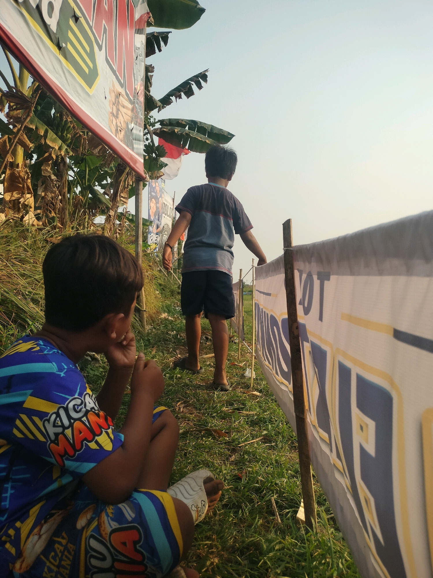 The image captures a scene at a grassy outdoor location, possibly near a local event or gathering, given the presence of banners. Two young boys are the primary subjects. The boy in the foreground is seated with his back to the camera, facing towards the right. He is wearing a brightly colored blue t-shirt with yellow, orange, and light blue graphics, and matching blue shorts with similar patterns. His left hand is near his face, and his posture suggests contemplation or observation.
A second boy is standing further away, also with his back to the camera, facing towards the right and slightly upwards. He is wearing a grey t-shirt with a dark stripe across the chest and dark shorts. He appears to be looking at something in the distance or at the sky.
Several banners are visible. A large, partially obscured banner with red and yellow lettering is in the upper left of the frame, behind banana trees. A white banner with blue and yellow lettering is stretched out horizontally on the right side of the image, supported by wooden poles. The lettering on this banner is partially visible and appears to be in Indonesian. The environment is characterized by dry grass, some green grass, and banana trees, suggesting a tropical or subtropical climate. The lighting indicates it is either early morning or late afternoon, with a pale sky. The presence of banners and what looks like a community setting might indicate a local festival or event in Kalisari, Indonesia.