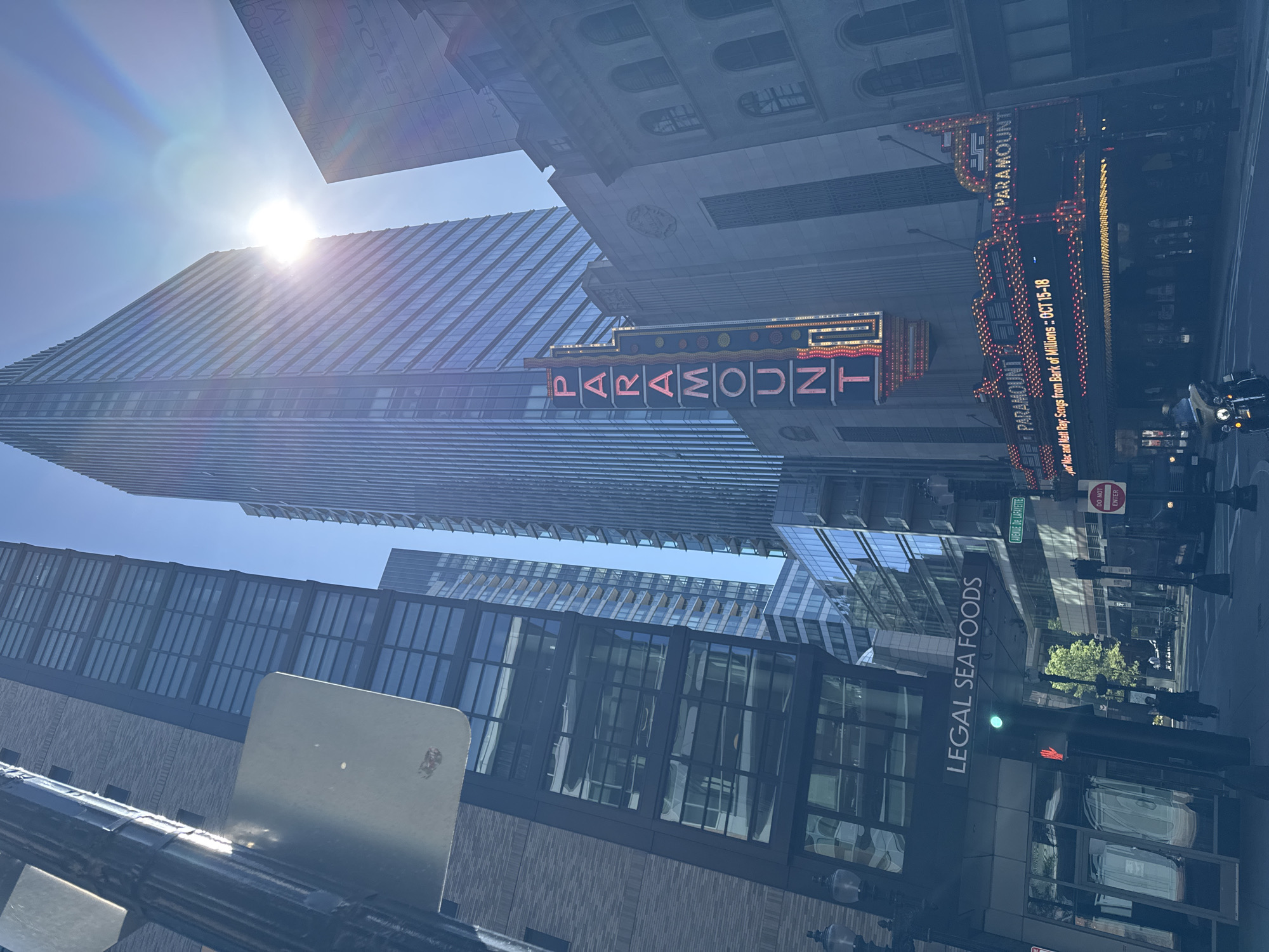 The image captures a vibrant, sunlit urban street scene, likely taken during the day, looking upwards towards tall buildings. The bright sun is positioned near the top of the frame, creating a prominent lens flare and illuminating a modern glass skyscraper that reflects the clear blue sky.
In the mid-ground, a classic theater marquee for the "PARAMOUNT" is prominently displayed, featuring its name in large, illuminated red letters against a dark background, accented with numerous yellow lights. Below this, a digital marquee advertises a performance: "Mac and Matt Ray. Songs from Bark of Millions :: OCT 15-18". Further up, parts of other building signs are visible, including "NORMANDIE BALLROOM" and "BIJOU".
At street level, a large vertical sign for "LEGAL SEA FOODS" is visible on the side of a building. Street infrastructure, such as lampposts, traffic lights, and street signs, are also present. One street sign clearly indicates "AVENUE DE LAFAYETTE", and a "DO NOT ENTER" sign is also visible. The overall impression is a bustling city environment on a clear, bright day, with no discernible people but clear signs of urban activity. The combination of the Paramount Theatre, Legal Sea Foods, and Avenue de Lafayette strongly indicates the location is Boston, Massachusetts, United States.