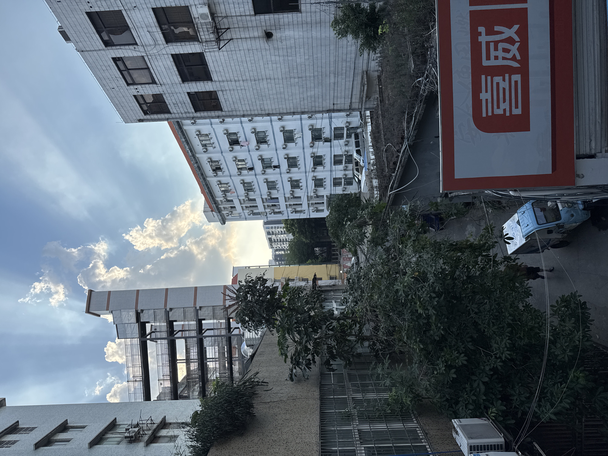 The image shows an urban scene in what appears to be Tianhe District, China, characterized by densely packed buildings and a bustling street-level environment. The architecture includes both older and newer structures, with white or light-colored facades. One building has numerous window-mounted air conditioning units. The sky is partly cloudy with visible sunshine. Street level is partially obscured by trees, but a small blue vehicle is observed near a building sign with red Chinese lettering. People are present on the sidewalk near the vehicle, although their actions and interactions are not explicitly clear. Electrical lines are visible overhead. The scene evokes a sense of urban density and daily life in a Chinese city.
