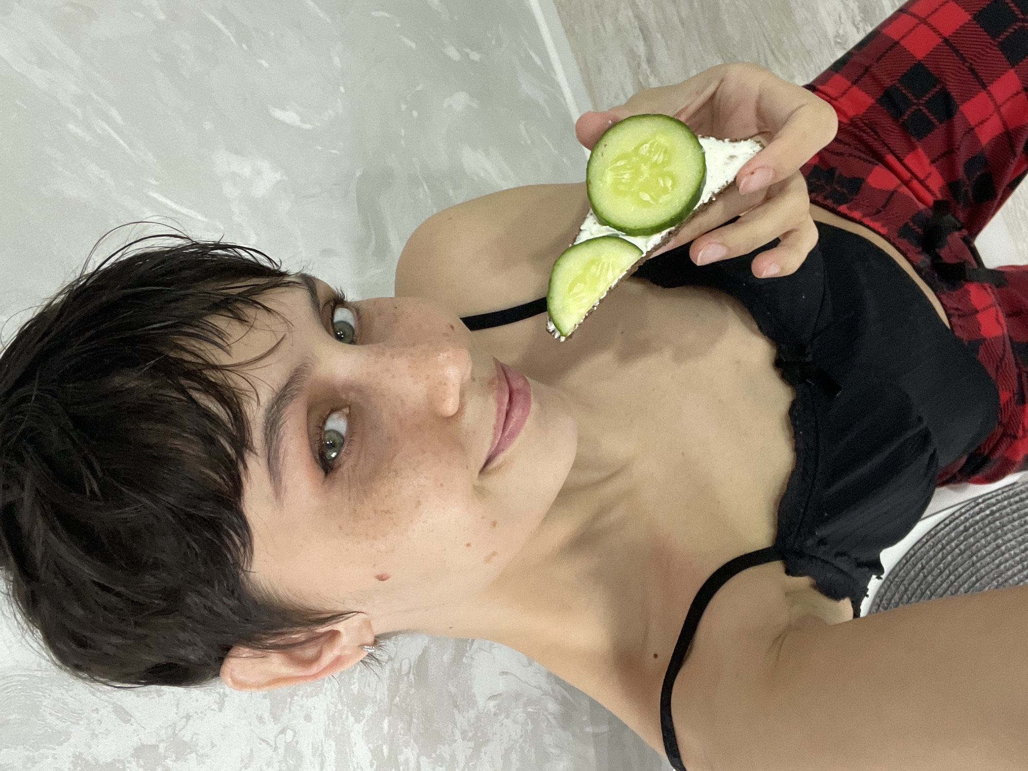 A woman with short dark hair is lying down, holding a sandwich with cream cheese and cucumber slices. She is wearing a black top with thin straps and a red and black plaid shirt. She has freckles on her face and an earring in her left ear. The background appears to be a light-colored wall or floor. The photograph was not taken in Izhevsk, Russia.