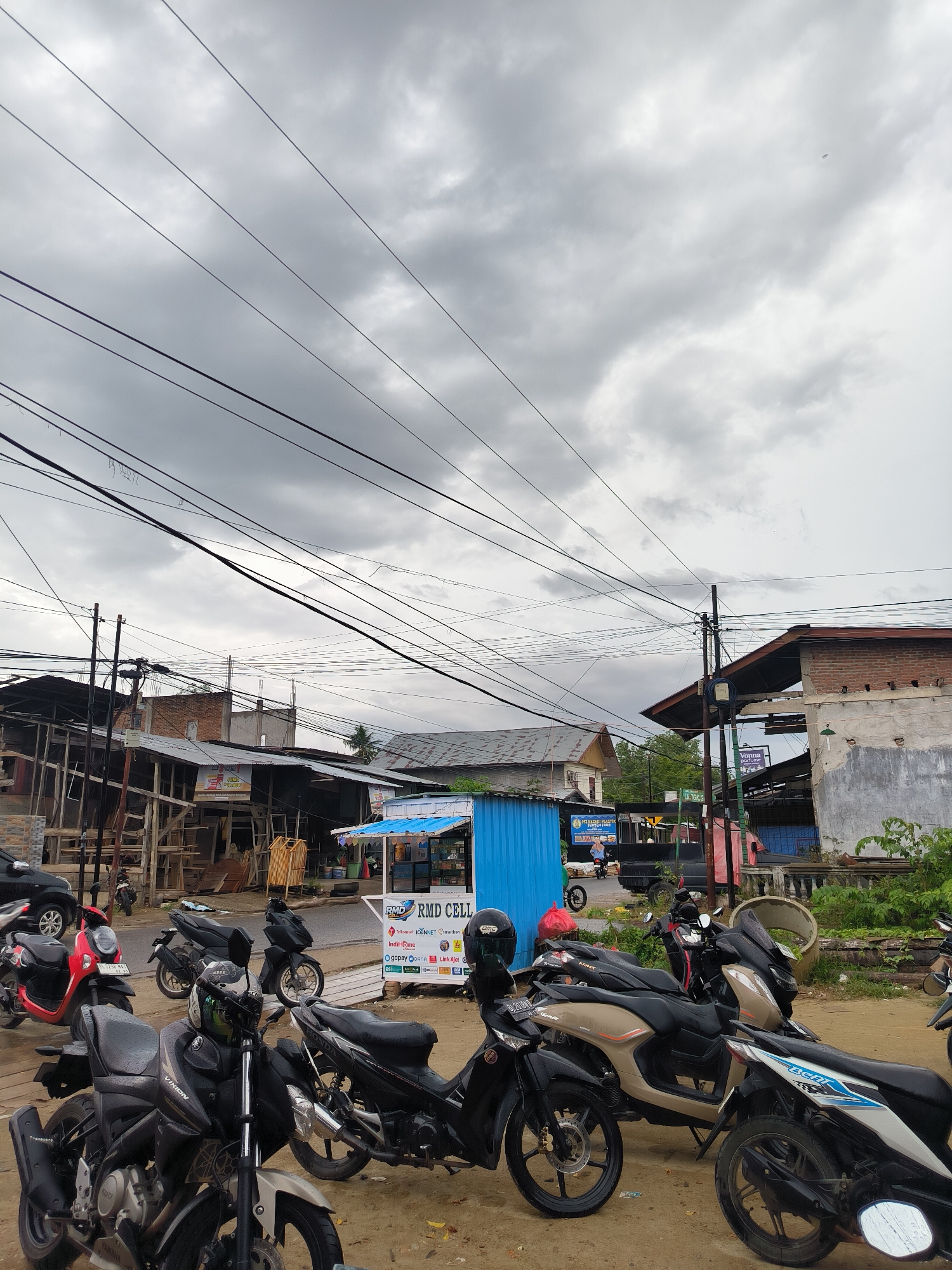 This media file captures a busy street scene in Lhokseumawe, Indonesia, under a heavily overcast sky. The weather appears cloudy and grey, suggesting either a pre-rain condition or a generally dull day, with soft, diffused lighting.
The foreground and midground are dominated by numerous parked motorcycles and scooters, common modes of transport in the region. Brands like "VIXION" (Yamaha) and "BEAT" (Honda) are clearly visible, with one scooter bearing the license plate "BL 3296 MAT". Helmets rest on some of the parked bikes, indicating recent use.
In the midground, a vibrant blue corrugated metal stall with a white awning stands out, identified by the sign "RMD CELL". This stall advertises various mobile and internet services including "Telkomsel", "ICONNET", "Smartfren", "IndiHome", and digital payment options like "gopay", "DANA", and "Link Aja!". Nearby, a "Vonna parfume" sign is also visible on another building. Further left, another blue stall or structure displays partial text mentioning "HPM SERBA PASTA" and "FRESH & FROZEN FOOD".
The background features a mix of buildings, some appearing aged, some under construction with exposed wooden frames and corrugated metal roofs. A dense network of overhead power lines and utility cables crisscrosses the sky, emanating from several utility poles, a characteristic sight in many Indonesian urban and semi-urban areas. A partial street sign "LR.TGK." can be seen. A black pickup truck and a few individuals are visible in the background, hinting at daily life and commerce. The ground is largely unpaved, appearing as dirt or gravel with some visible puddles, further indicating a local, perhaps less developed, street. The overall scene depicts a functional, bustling local area.