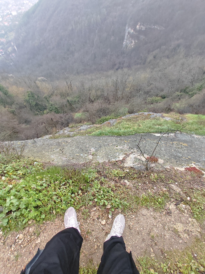 The image displays a downward-angled perspective from a cliff edge on the Shumen Plateau in Shumen, Bulgaria. In the foreground, the lower legs and feet of an individual are visible, clad in black trousers and light-colored athletic shoes, positioned on a patch of earthy ground interspersed with sparse green vegetation. Immediately ahead, the terrain consists of weathered grey rock and scattered green plant growth, leading to a sharp drop-off. The midground shows a steep descent into a heavily forested valley, where numerous deciduous trees appear bare, suggesting a winter or early spring season. In the upper-left background, a portion of the city of Shumen is discernible, featuring residential structures with reddish roofs, partially obscured by atmospheric haze or distance. The sky above is uniformly overcast, casting a diffused light over the landscape. No direct actions or interactions beyond the static presence of the individual's lower body are depicted.