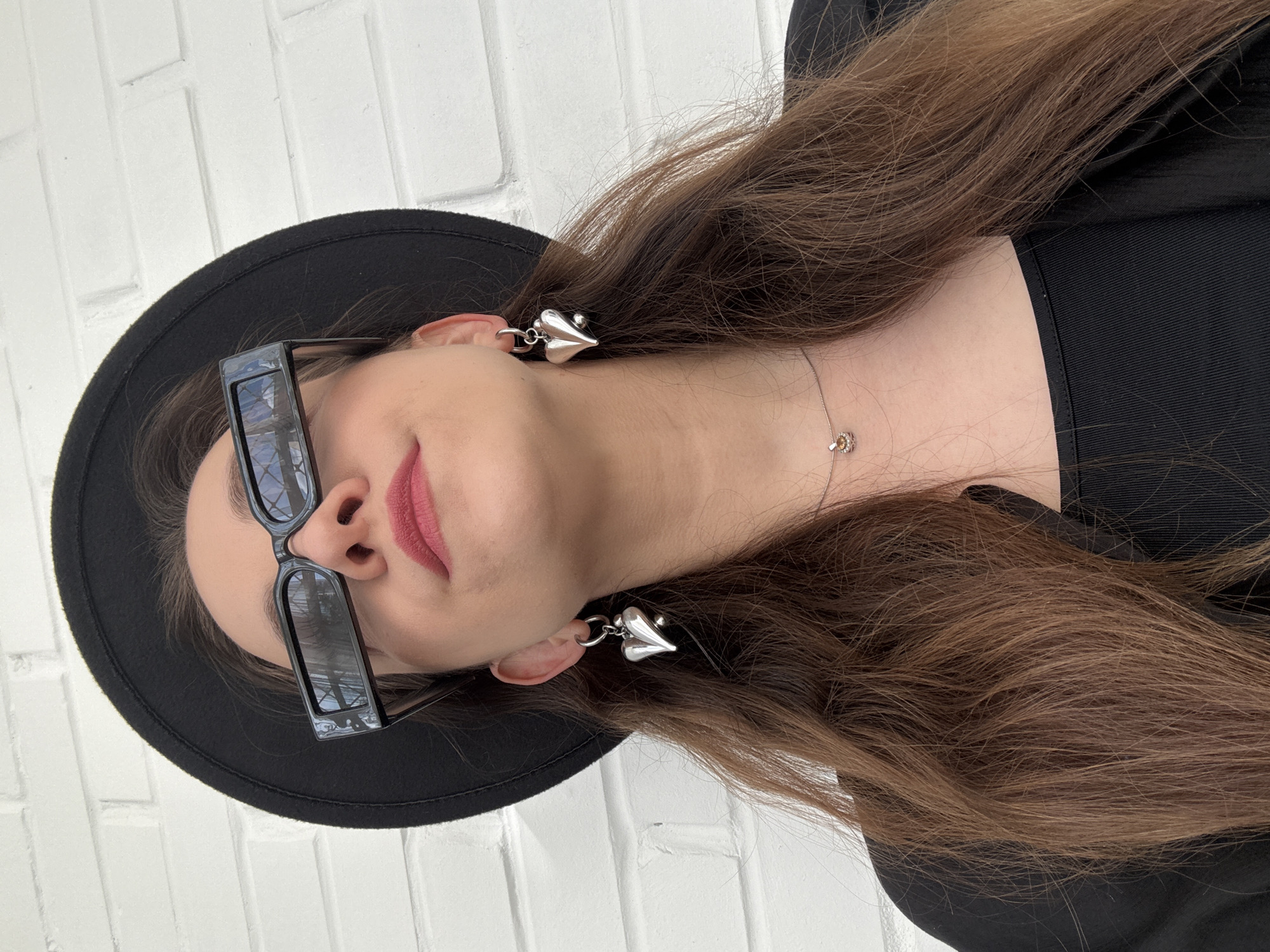 A person wearing a black hat and sunglasses is lying down on a white brick surface. They are wearing a black garment. Heart-shaped earrings dangle from their ears and a necklace adorns their neck. The location is an open outdoor space possibly located in the city of Chaikovsky, Russia.