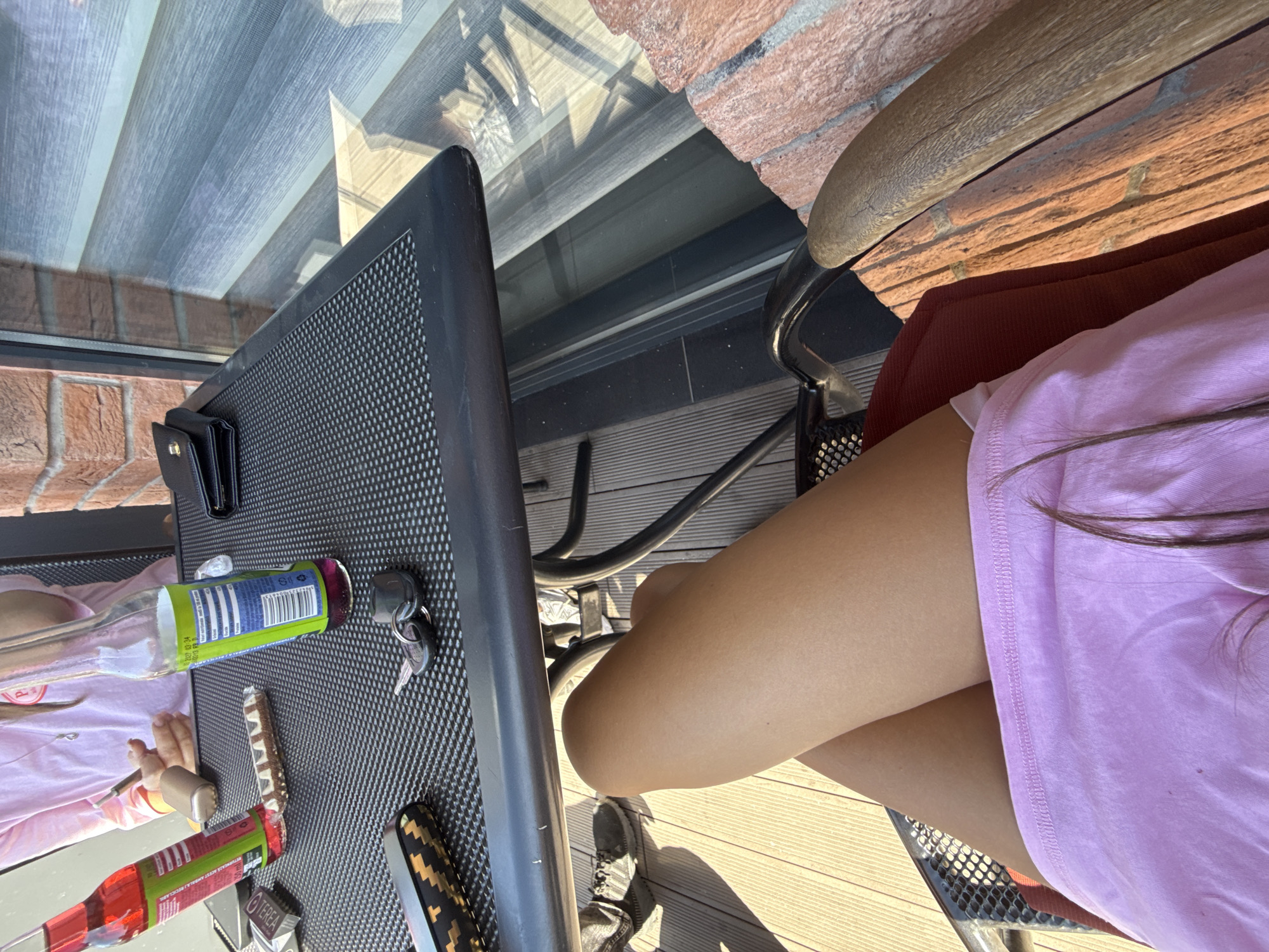 The image captures a close-up view of a person's legs, clad in a pink skirt, seated on a chair on a wooden deck. The scene appears to be an outdoor seating area, possibly a balcony or patio, with a table in the foreground. On the table are two bottles of beverages, a wallet, a chocolate bar, and a set of keys. The background shows a brick wall and a window with blinds. The sunlight suggests it is daytime. This setting is characteristic of a residential area in Comrat, Moldova, where such outdoor spaces are common for leisure and relaxation.