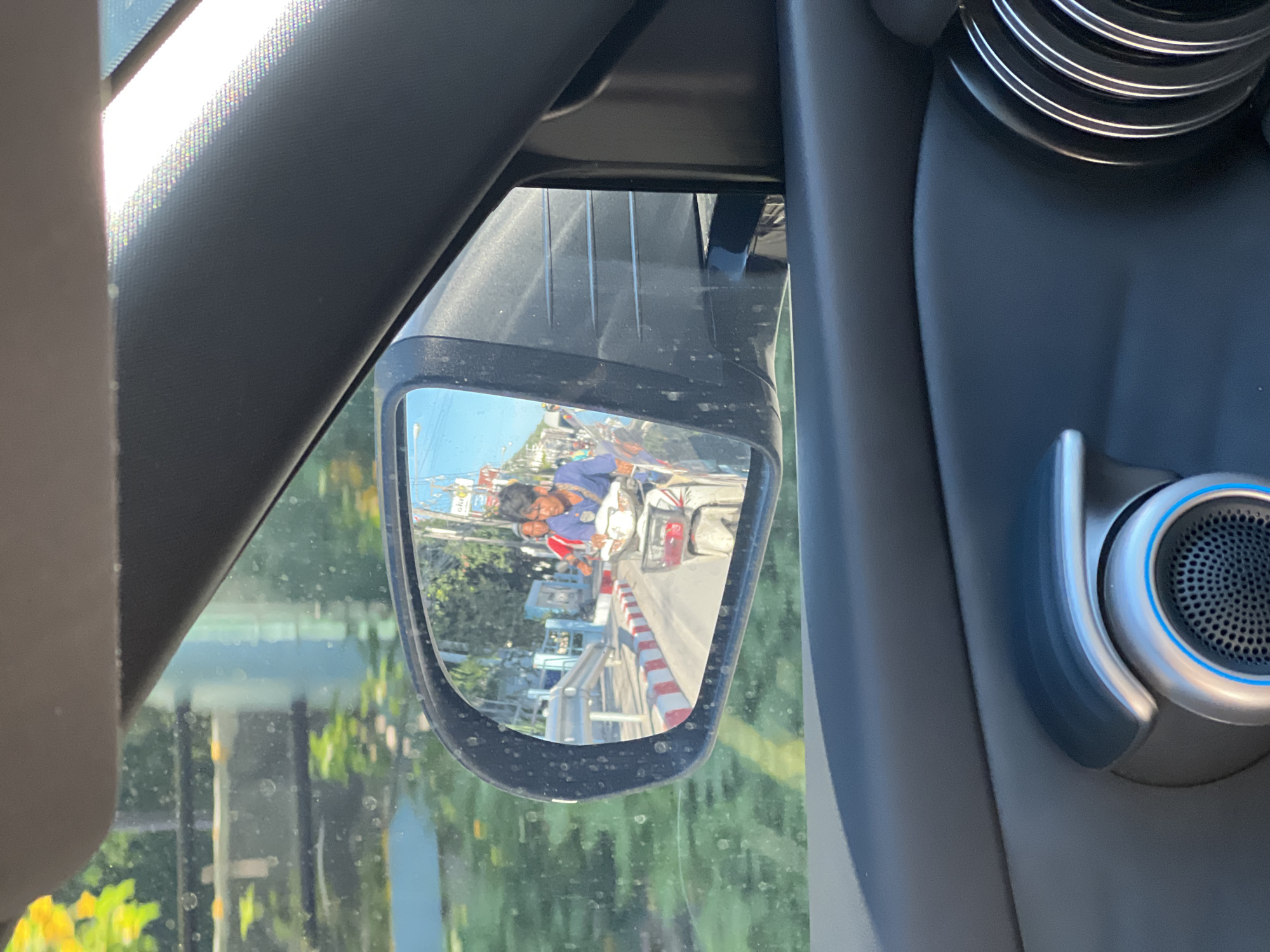 The image is taken from inside a car, looking out at the side-view mirror. The mirror reflects a street scene in Baan Mae Nam, Thailand. In the reflection, two men are riding a scooter. The driver, in a blue shirt, is looking forward. The passenger, in a red shirt, is looking to the side. The scooter has a basket on the front. The street appears to be paved with a red and white striped curb. There are buildings and trees in the background. The sky is clear and blue. The car interior is visible in the foreground, including part of the door panel and a speaker.