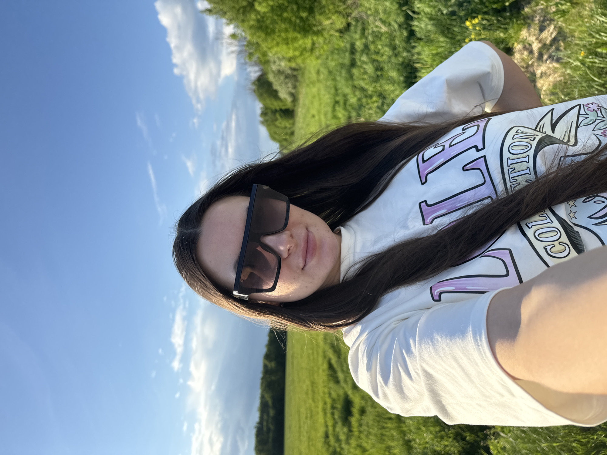 A young woman with long dark hair is taking a selfie outdoors. She is wearing sunglasses and a white t-shirt with a graphic print. The background consists of a blue sky with scattered clouds and a green field with trees in the distance. The location is Bolshiye Vyazyomy, Russia.