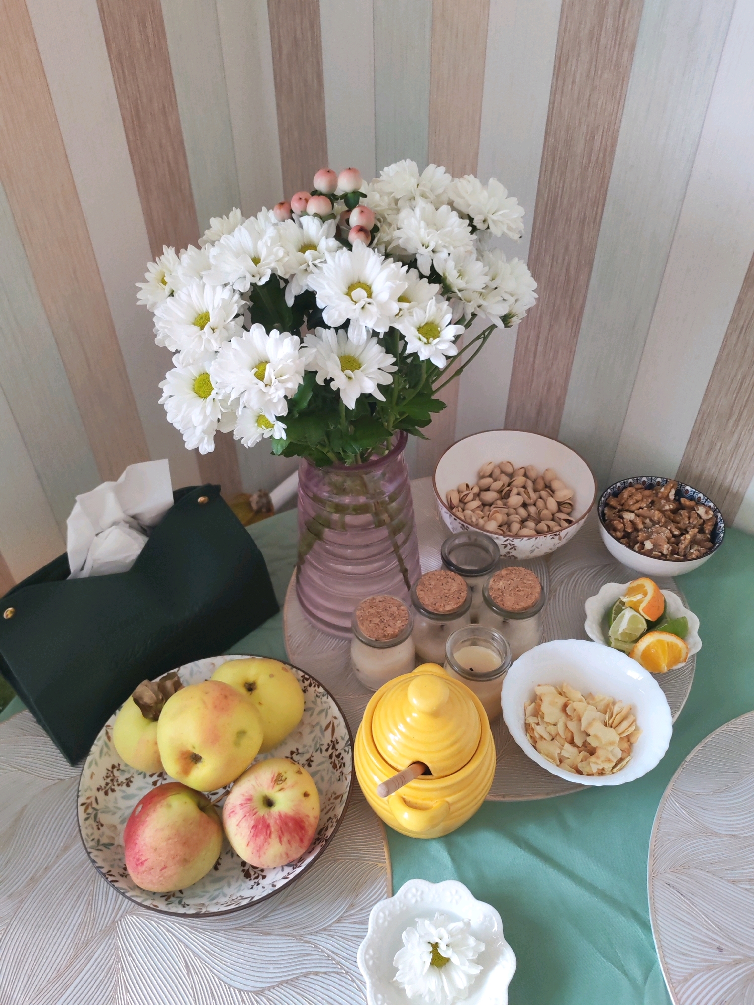 The image depicts a tabletop spread with various food items and decorations, set against a backdrop of a striped wall. The scene appears to be indoors, likely in a home or a cafe.
In the center of the arrangement, a bouquet of white chrysanthemums with pink berry accents is displayed in a pink glass vase. To the left of the flowers, a dark green tissue box holder contains white tissues.
Arranged on a patterned plate are five apples, some with red blush and others primarily yellow. Beside the apples, a bright yellow ceramic honey pot with a wooden dipper sits on a pale green tablecloth.
Several small glass jars with cork stoppers are lined up, some appearing to contain candles. A white bowl filled with pistachios and another with walnuts are also visible. A small white bowl holds slices of orange and lime. A white oval bowl contains what appear to be toasted almond flakes.
In the foreground, a small white decorative dish holds a single white flower. The table is covered with a pale green tablecloth, and decorative placemats with a leaf-like pattern are placed under some of the bowls.
The overall impression is of a breakfast or snack setup, with a focus on fresh produce and healthy snacks.
There is no information in the image to suggest the location is the city of Izhevsk, Russia.