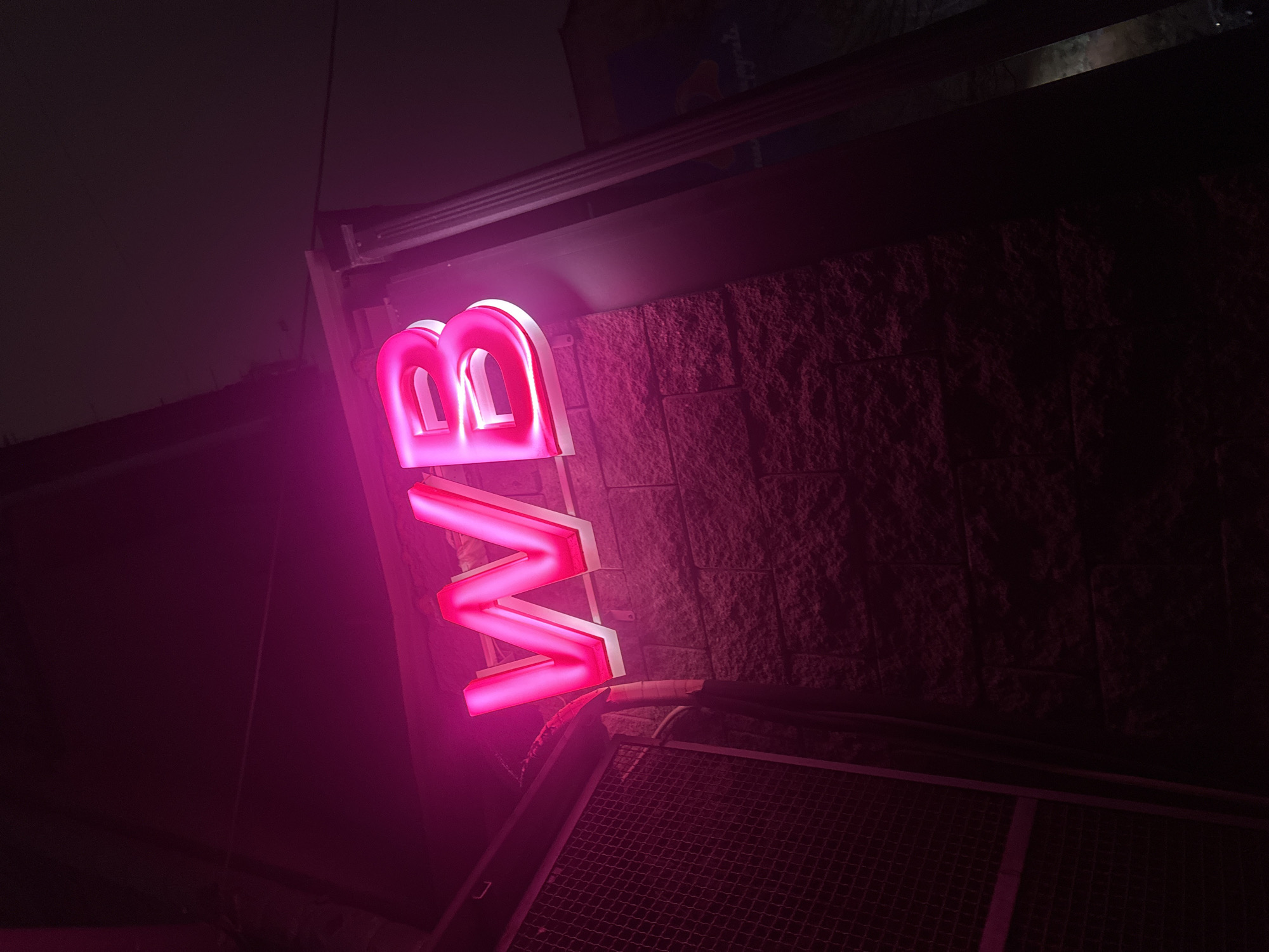 Here's a detailed description of the image:
**Scene:** Nighttime view of a building exterior.
**Objects:**
* A bright pink neon sign with the letters "VB" vertically stacked is prominent against a brick wall.
* A metal grate or vent-like structure is located below the sign.
* The building has an overhang or ledge above the sign, with some visible structural elements.
**Environment:**
* The overall scene is dark, with the neon sign providing the primary source of light.
* A dark building wall behind the sign.
**Actions/Interactions:** Not applicable, as the image is static and does not capture any actions.