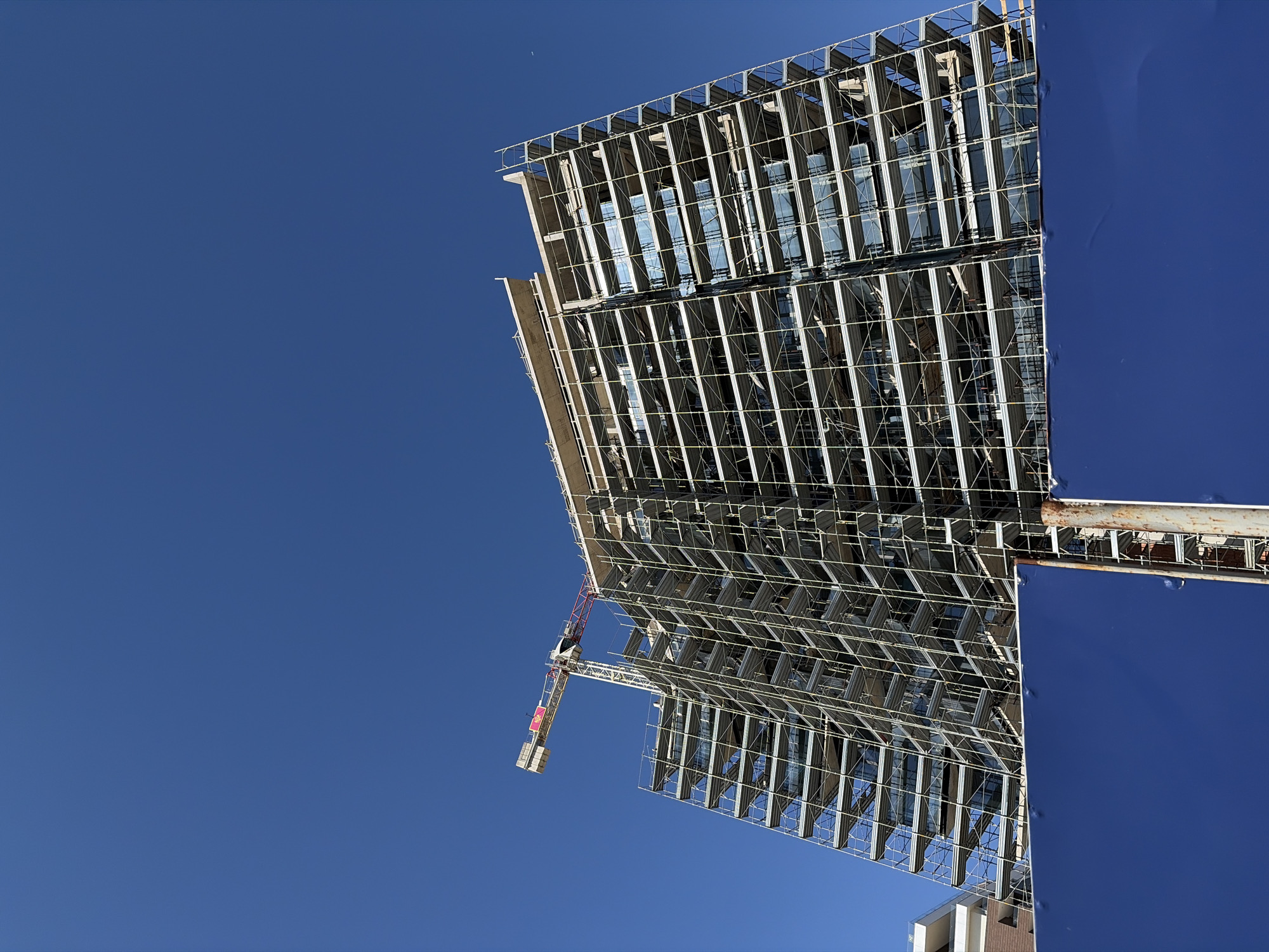 The image shows a building under construction in Budva, Montenegro. The building's steel framework is visible, with sections of glass windows in place. A crane is attached to the building's structure. The sky is clear blue. A blue screen partially covers the building's side.