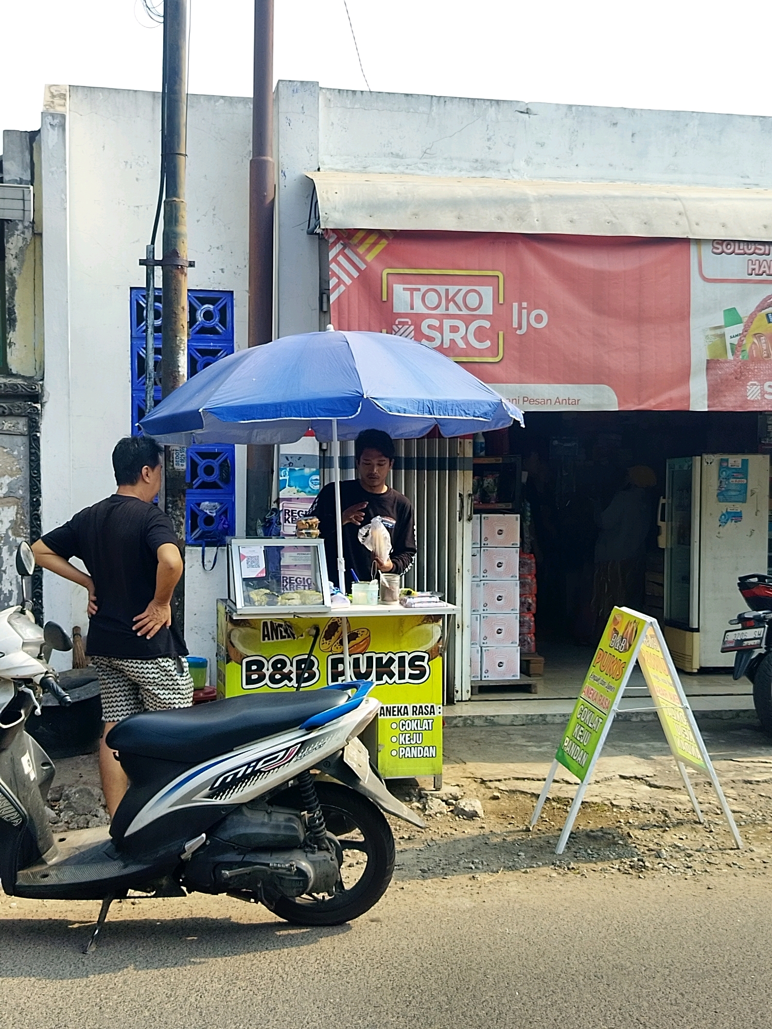 In Mojokerto, Indonesia, a street vendor stands behind a small food cart shaded by a blue umbrella, serving a customer. The vendor, wearing a black long-sleeved shirt, holds a bag of food. The customer, in a black t-shirt and patterned shorts, faces the cart with hands on hips. A white and blue scooter is parked in the foreground. Behind the cart, a storefront with a red banner that reads "TOKO SRC IJO" is visible, along with stacked boxes and a refrigerator. A yellow A-frame sign with text about food flavors stands on the sidewalk. The scene is set along a paved street with a cracked pavement.