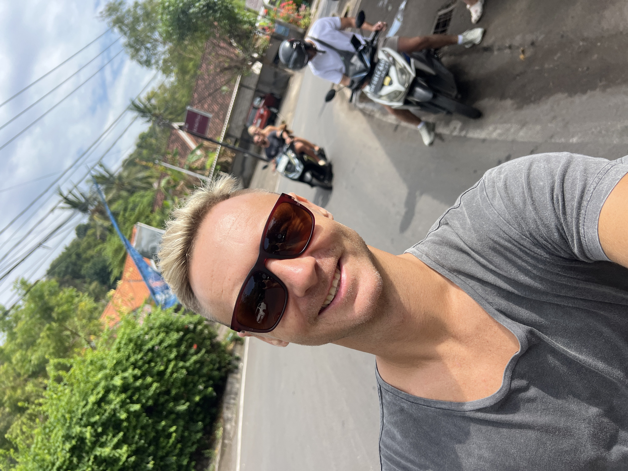 A man with blond hair and sunglasses smiles at the camera, taking a selfie while riding a motorbike. He wears a grey t-shirt. Behind him, two other motorbikes are visible on a paved road lined with trees and buildings. The scene appears to be a street in Abang, Indonesia, with lush green hills in the background. The sky is cloudy.