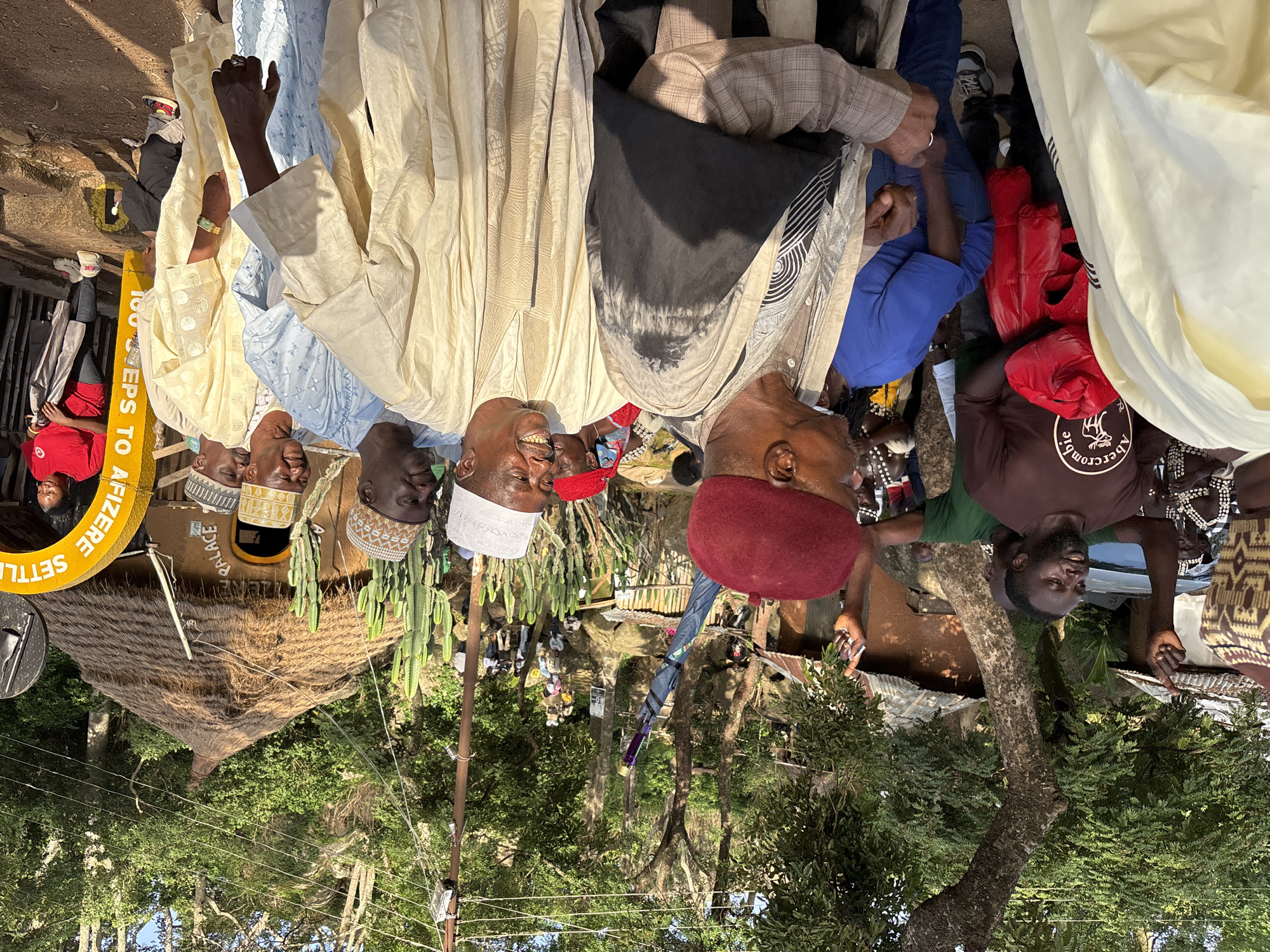 This media file captures a gathering of men, likely in Jos, Nigeria. The subjects are predominantly adults, dressed in traditional African attire, including flowing robes and patterned caps. Some individuals are smiling broadly, suggesting a positive and convivial atmosphere.
The scene appears to be outdoors, possibly at a community event or a traditional ceremony. A thatched-roof structure, resembling a hut or small dwelling, is visible in the background, along with dense green foliage and trees. Several tall, columnar cacti are also present, adding a distinctive visual element to the setting. Some men are holding umbrellas, one of which is long and dark with a purple tip, possibly for shade or as a ceremonial item.
One man wears a red cap, while others sport white or patterned caps. The presence of a yellow circular sign with some text suggests an organized location or event. The lighting indicates it is daytime, likely late morning or afternoon, with bright sunlight casting shadows. The overall mood seems to be one of community and celebration.
Visible text includes "PALACE" on a structure and "SETTLE" and "100 STEPS TO AF..." on the yellow sign. The image is oriented sideways.