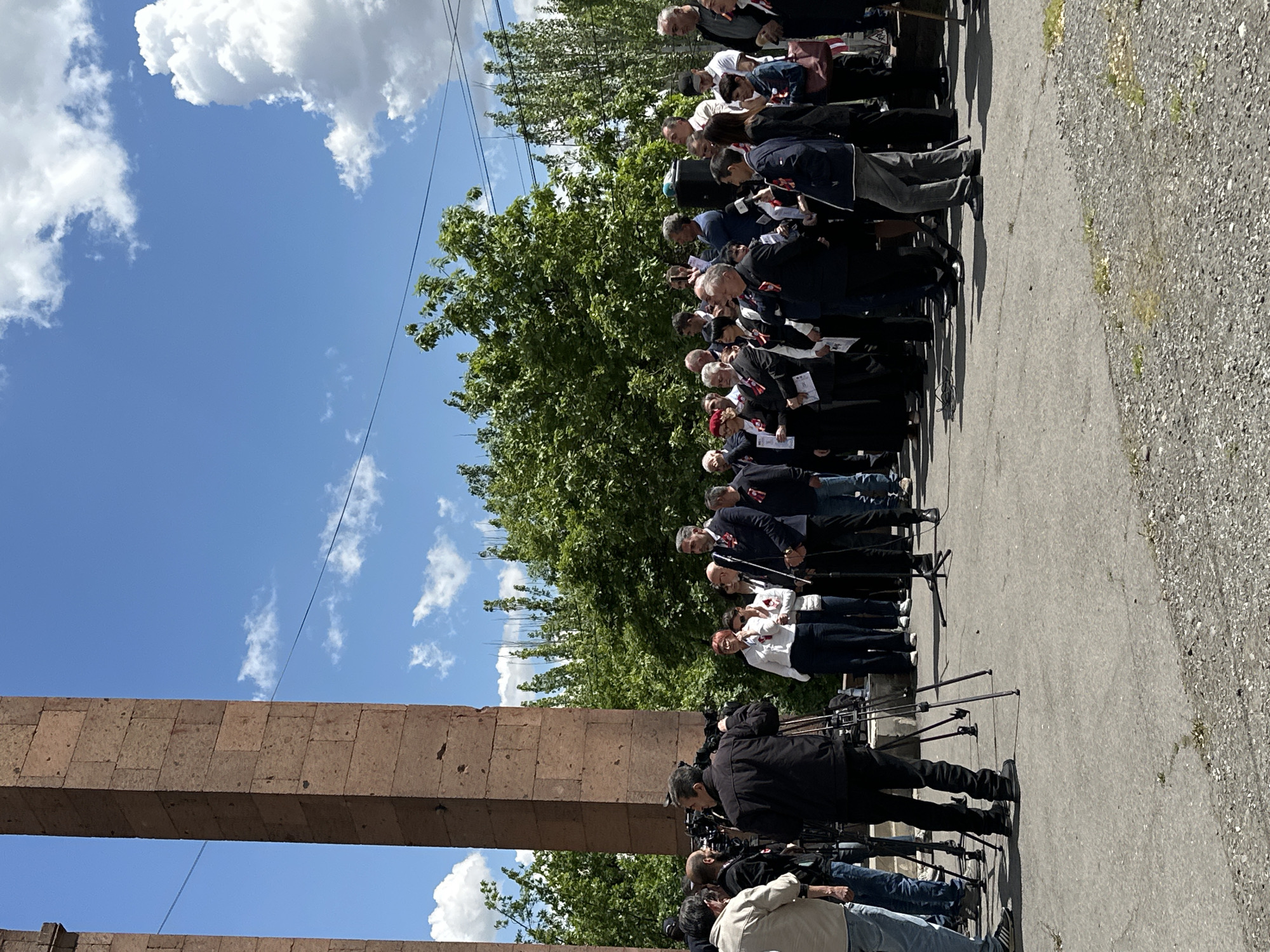 The image shows a group of people standing in a line in Yerevan, Armenia. They are standing on a paved surface next to a large stone structure. Many individuals appear to be partaking in a formal occasion with a contingent of media in attendance. The sky is visible in the background and is blue with white clouds. A tree is visible behind the line of people. The time of day appears to be daytime.