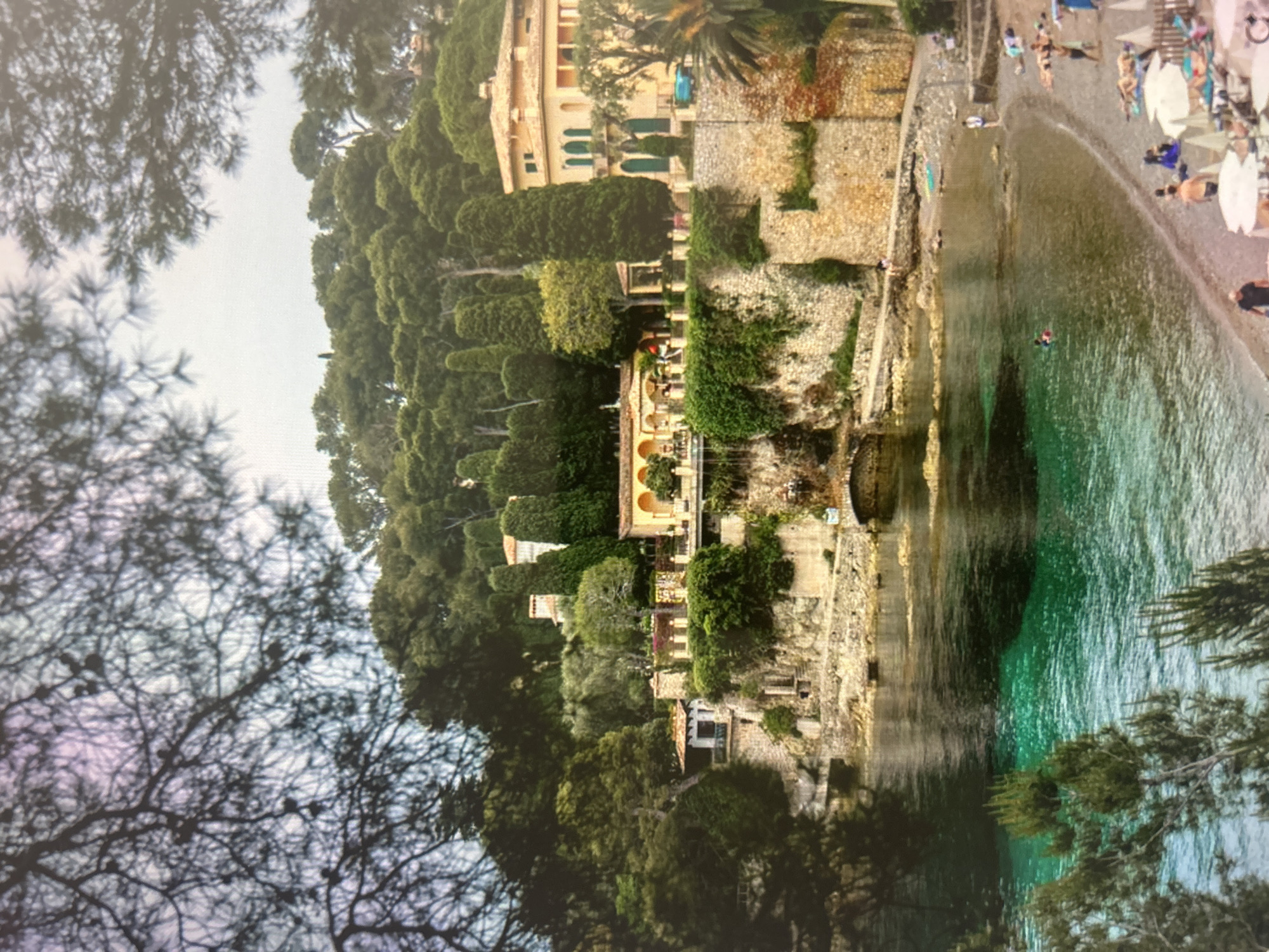 This image captures a picturesque Mediterranean coastal scene, likely a resort or village, on a bright day. The perspective is slightly elevated, looking down towards a bay, and is partially framed by silhouetted tree branches in the foreground.
On the left, a steep hillside is densely covered with lush green vegetation, dominated by tall, slender evergreen trees and various other broadleaf trees. Several buildings with light yellow or ochre-colored facades and terracotta roofs are nestled into the hillside. These structures exhibit architectural details such as green-shuttered windows, arched balconies, and stone walls.
The center and right side of the frame feature a beautiful, clear bay with water exhibiting striking shades of turquoise and emerald green. A crescent-shaped sandy or pebbly beach curves along the coastline. Numerous people are visible on the beach, engaged in leisure activities. Many are relaxing on lounge chairs under light-colored beach umbrellas, while others are standing or strolling along the shore. Several individuals are seen swimming or wading in the pristine water, with one person distinctly visible in the clear sea. A small blue boat and another lighter-colored boat are moored near the beach.
The scene conveys a sense of relaxation and natural beauty, typical of a holiday destination. The time of day appears to be mid-day, with bright, possibly hazy, skies. There is no visible text in the image.