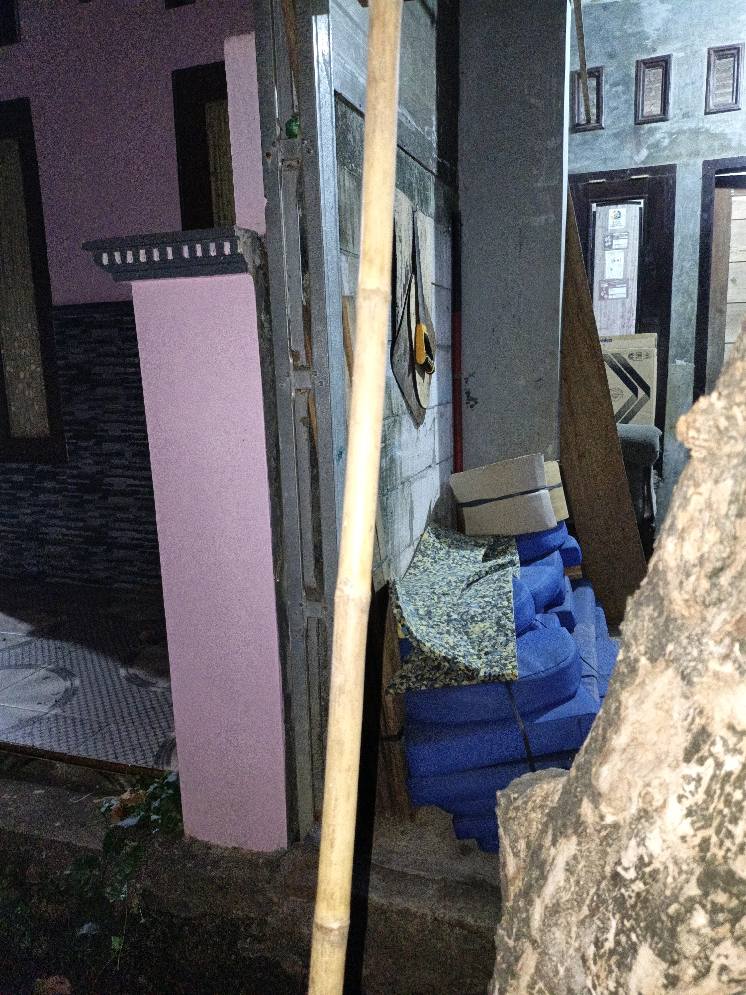 In Cikal, Indonesia, a snapshot captures the exterior of a building. A tall, pink pillar stands prominently to the left, adorned with a dark grey decorative ledge featuring rectangular cutouts. To the right of the pillar, a thin, upright bamboo pole leans against a metal frame, which is part of the building's structure. Further to the right, a stack of blue cushions is visible, partially covered by a piece of patterned foam. Behind the cushions, a large, diagonally placed wooden plank leans against the wall. The wall itself is textured and grey, with three small, dark-framed windows positioned above two dark-colored doors. One of the doors has a sign with text and images attached to it. In the foreground, a rough, textured tree trunk occupies the right side of the frame. The ground is a mix of dark soil, greenery, and patterned paving stones to the left. The lighting suggests it is either dusk or a dimly lit area.