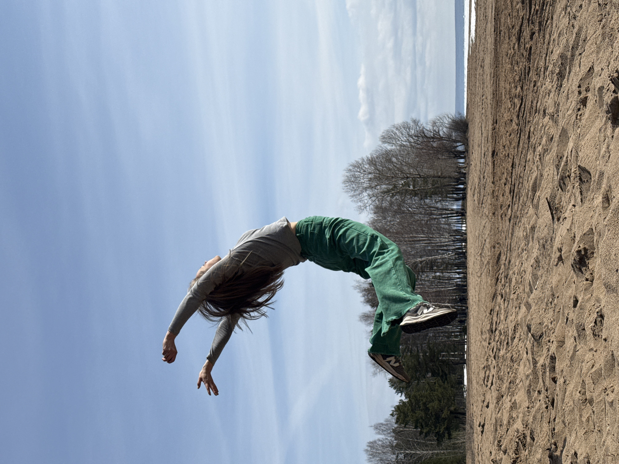 The image shows a person performing a backbend on a beach. The person is wearing a grey long-sleeved shirt and green pants and is barefoot or wearing sneakers. The beach appears to be sandy. In the background, there are trees. The sky is partially cloudy.
It is not possible to confirm from this image that the location is Noviy, Russia.