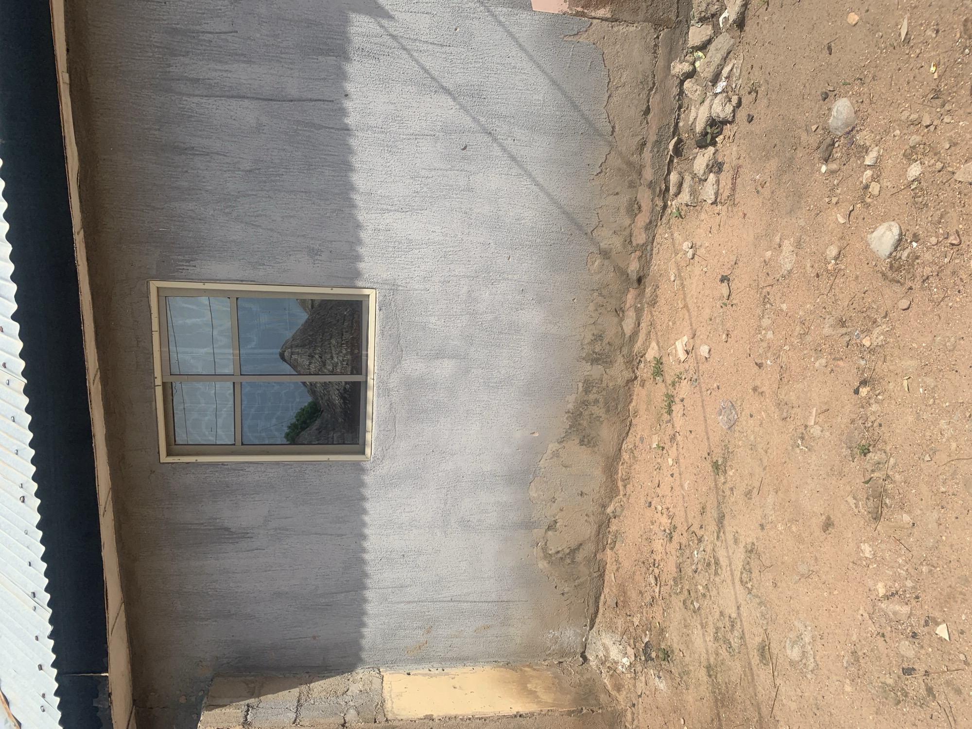 This image depicts an exterior view of a building under construction in Jalingo, Nigeria. The primary subject is a rough, gray stucco wall with a small, aluminum-framed window. The window is divided into four panes and reflects a thatched roof structure and some greenery, suggesting the presence of other buildings or natural elements nearby. To the left of the wall, a section of a corrugated metal roof is visible, indicating the building is at some stage of completion. The ground in front of the wall is a mix of dry, sandy soil, small stones, and some scattered debris. The lighting suggests it is daytime, with sunlight casting shadows on the wall. The overall scene conveys a sense of a developing or unfinished construction site in a potentially arid or rural environment. There is no visible text in the image.
