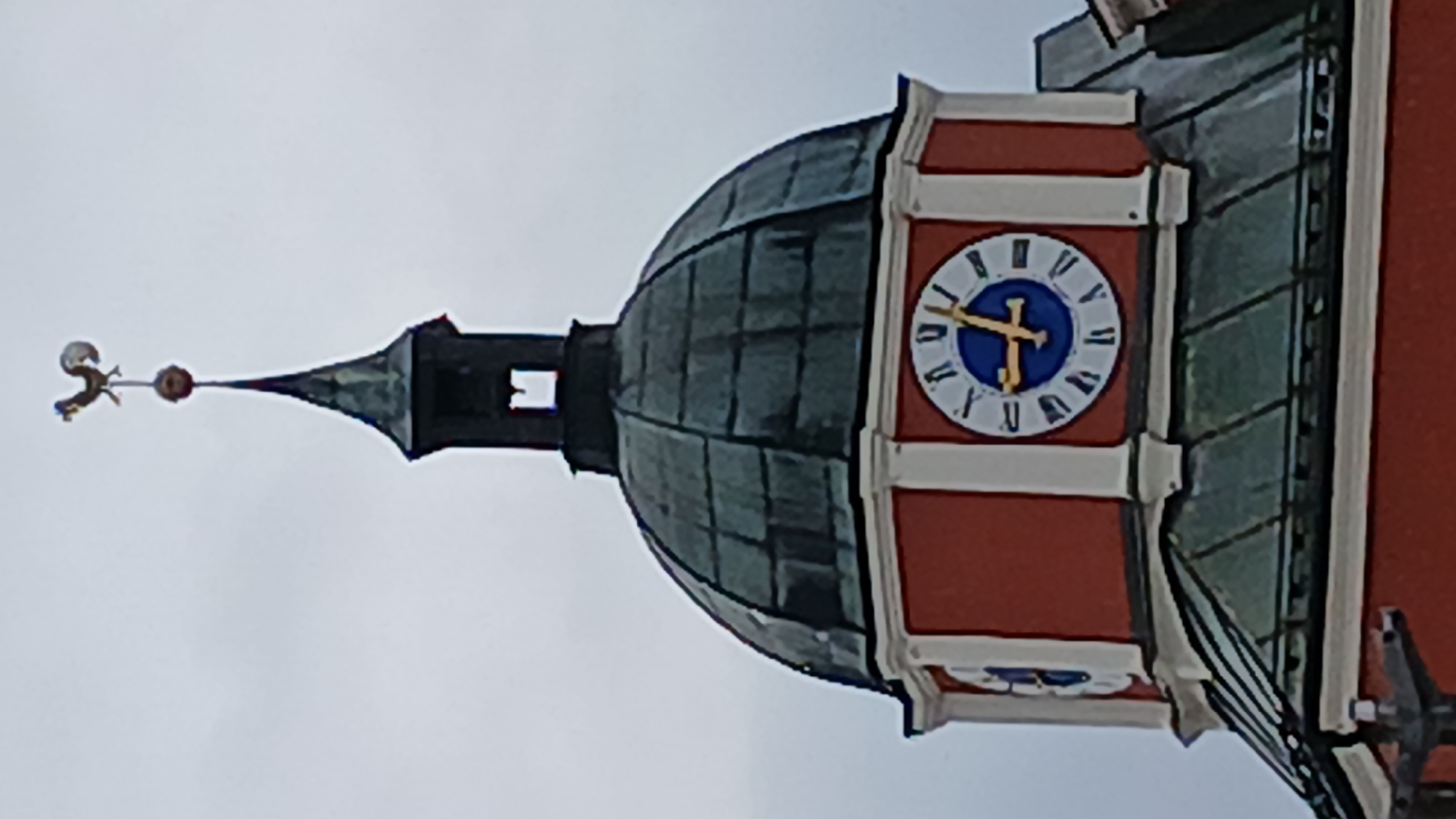 The image shows the upper section of the bell tower of the St. Jakobus church in Pfarrkirchen, Germany. The tower is topped with a weather vane shaped like a rooster. Below the weather vane is a dark spire, and then a red octagonal section featuring two clock faces. The clock faces have Roman numerals and blue and gold hands. The lower part of the image shows a green tiled roof section. The sky is overcast.
