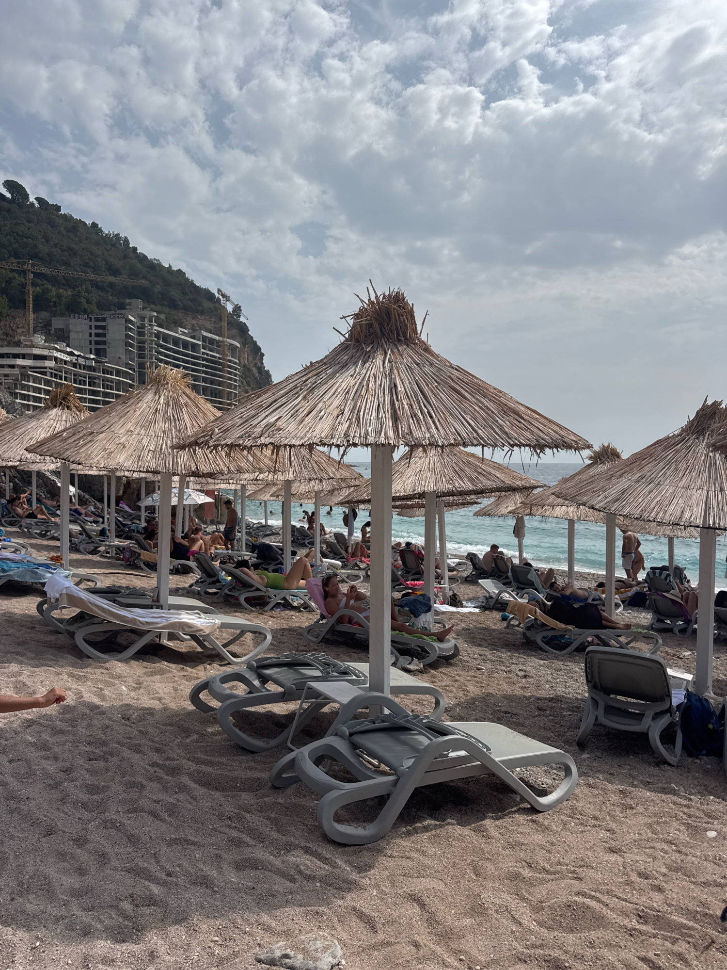 The image depicts a crowded beach scene in Mirište, Montenegro. Numerous thatched umbrellas provide shade for rows of empty and occupied lounge chairs on a gravelly beach. In the background, several unfinished apartment buildings under construction are visible on a hillside, along with construction cranes. People are scattered across the beach, some sitting on lounge chairs, others standing or walking. The sea is visible in the distance, with a few people swimming or wading in the water. The sky is cloudy.