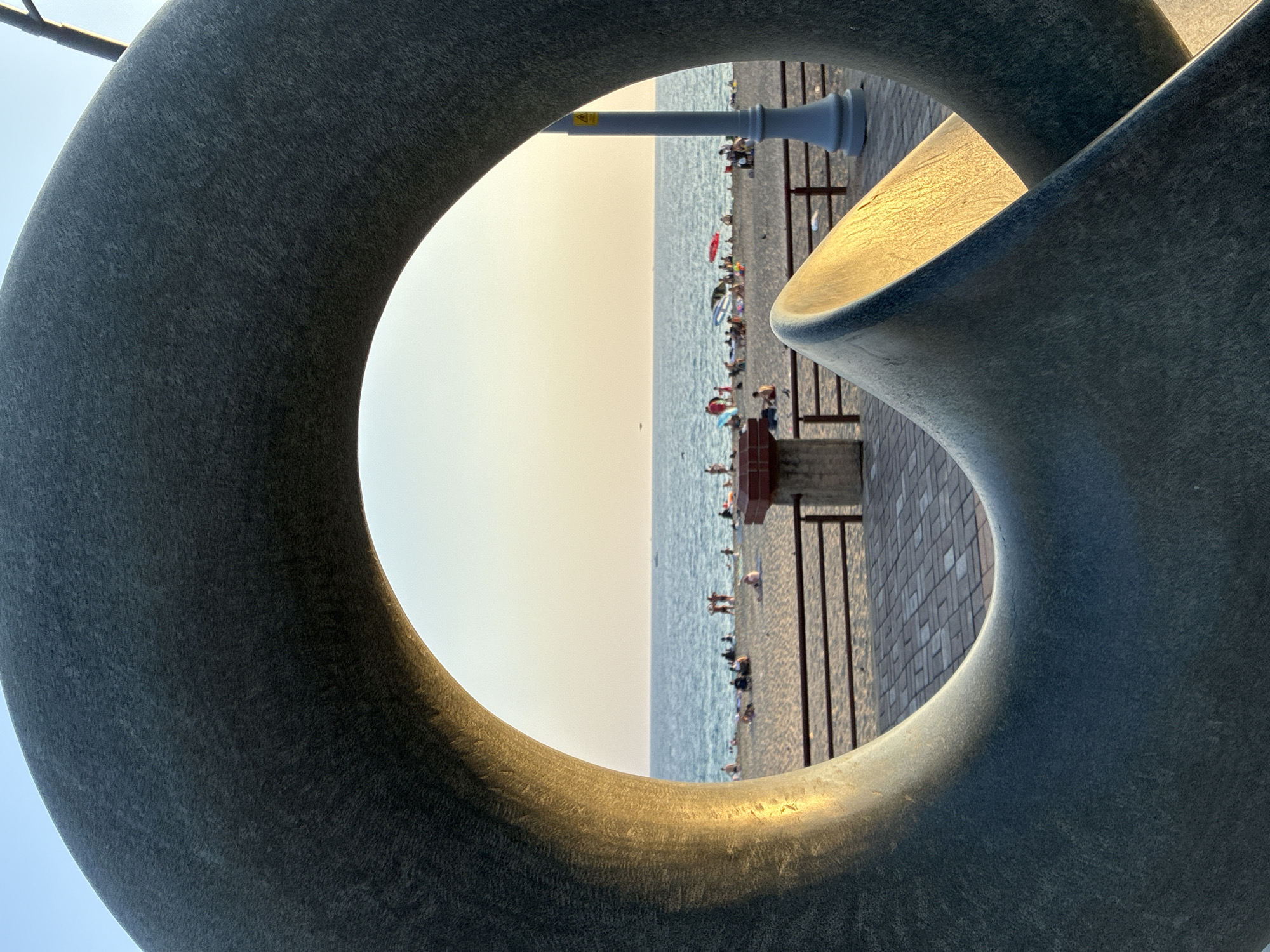 The image captures a view through a large, dark gray, circular sculpture, framing a beach scene in Alanya, Turkey. The sculpture, made of a textured material, occupies the foreground, with its curved surfaces catching the warm, golden light of what appears to be late afternoon or early evening. Through the circular opening of the sculpture, a sandy beach stretches out towards a calm, blue sea. Several people are visible on the beach, some sitting, some standing, and a few appear to be swimming or wading in the water. Beach umbrellas are dotted across the sand, providing pops of color. A boardwalk or paved area runs along the edge of the beach, with a railing separating it from the sand. In the background, the sky is a soft gradient of pale yellow and blue, suggesting a sunset or sunrise. A lamppost stands on the right side of the frame, partially visible through the sculpture. The overall impression is one of a peaceful, leisurely day at the beach.